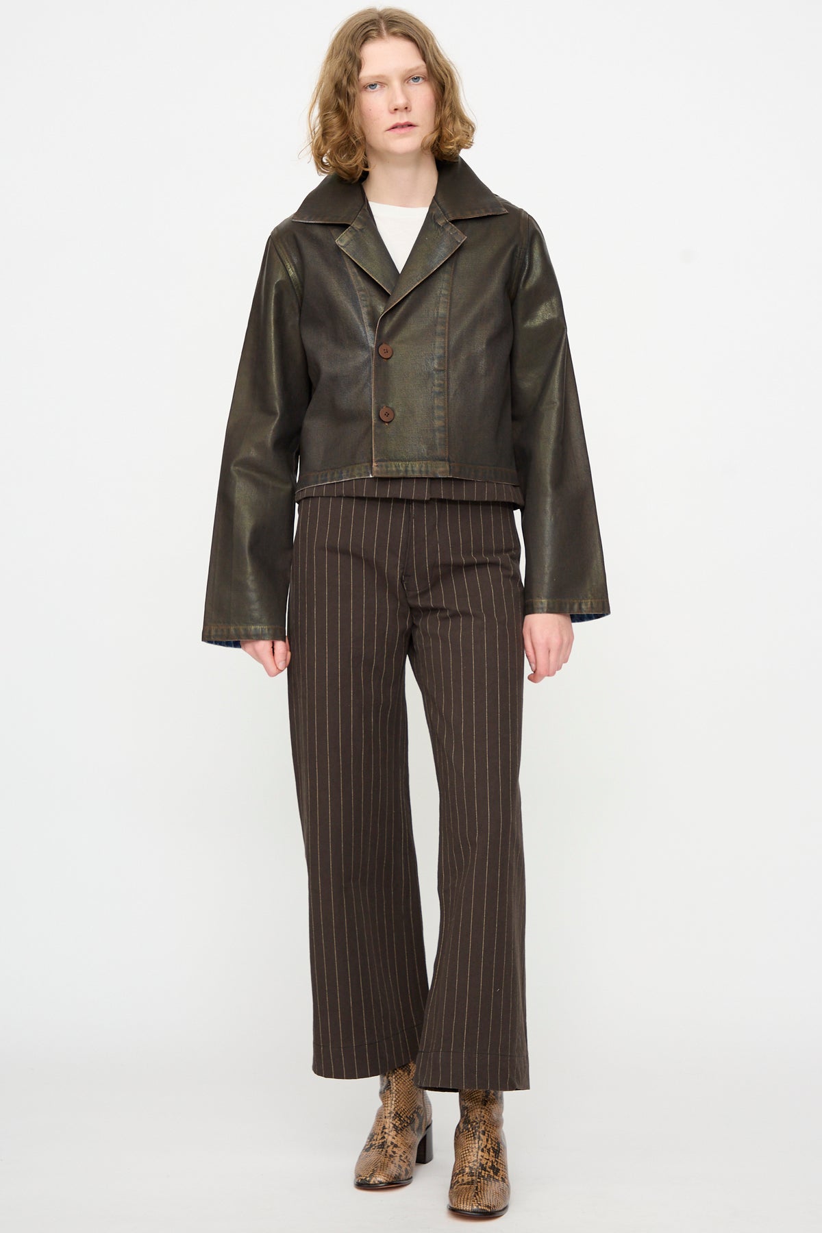 A person stands against a white background, wearing the Jesse Kamm Jones Jacket in Rich Brown Lacquer, along with brown pinstripe pants and patterned ankle boots. They have short wavy hair and a neutral expression.