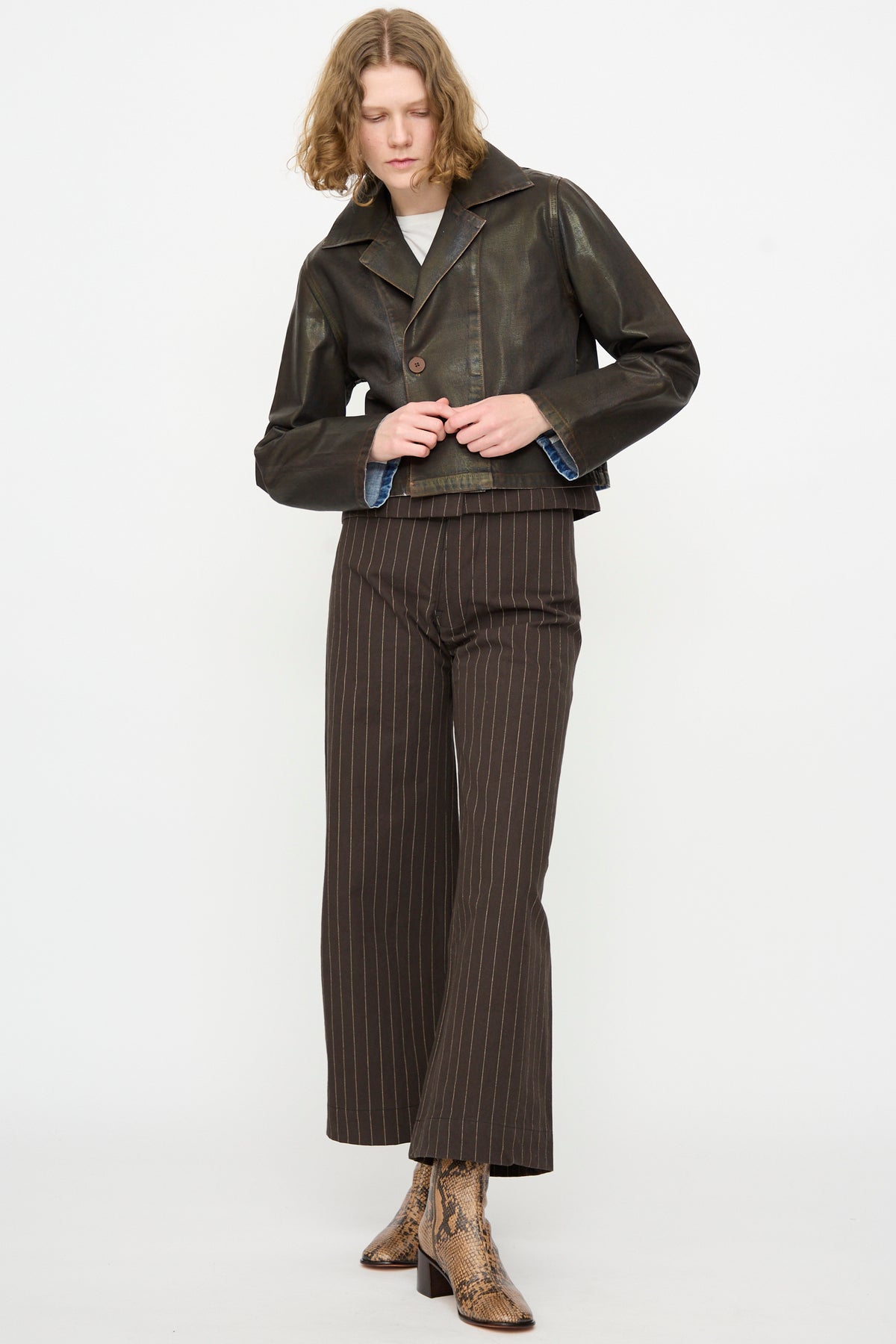 Person wearing the Jones Jacket in Rich Brown Lacquer by Jesse Kamm, paired with a white top, brown pinstripe wide-leg pants crafted from regenerative Japanese denim, and snakeskin boots, posing against a plain white background.