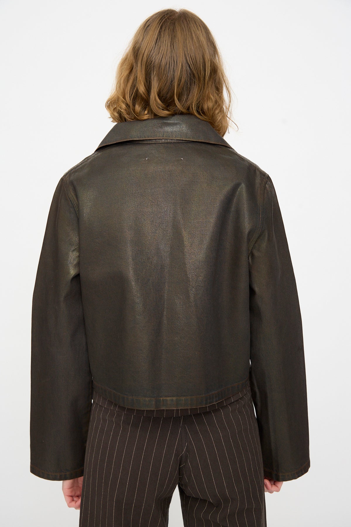 A person with shoulder-length light brown hair wears the Jesse Kamm Jones Jacket in Rich Brown Lacquer, crafted from regenerative Japanese denim, paired with pinstriped pants and shown from the back against a plain light background.