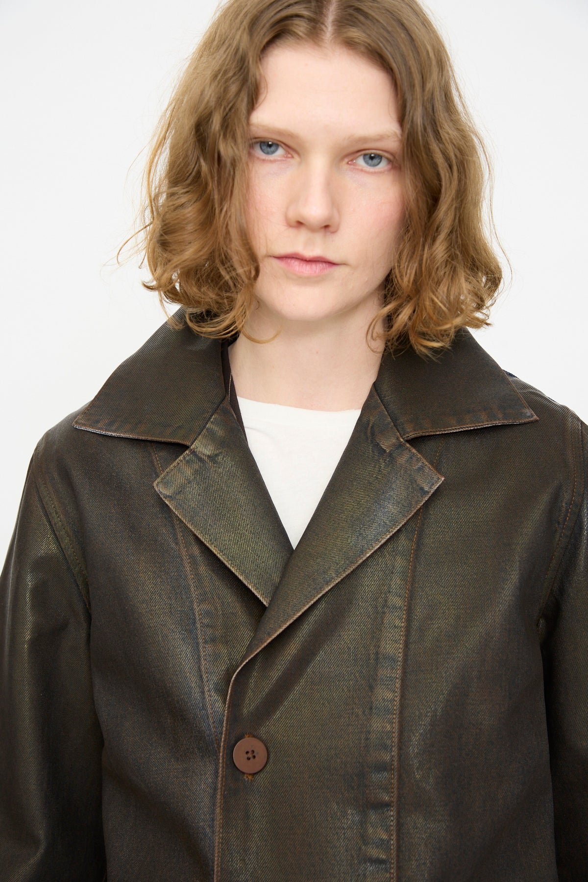 A person with shoulder-length wavy hair wears the Jesse Kamm Jones Jacket in Rich Brown Lacquer—a shiny, buttoned coat featuring hand-applied lacquer—over a light shirt, standing against a plain white background.