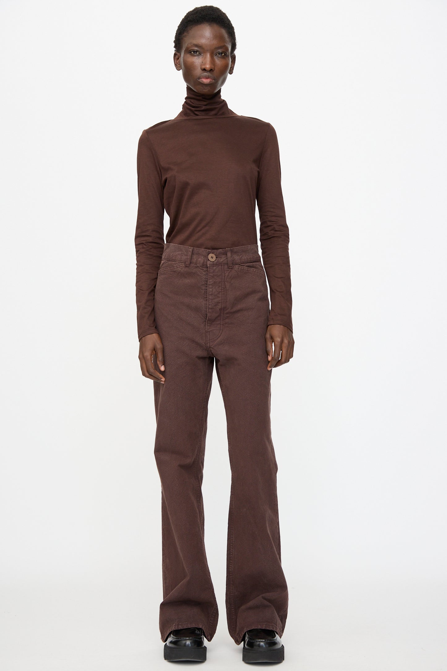 A person stands facing forward wearing Jesse Kamm Organic Canvas Les Deux Pants in Raisin, a brown turtleneck, and black shoes against a plain white background.
