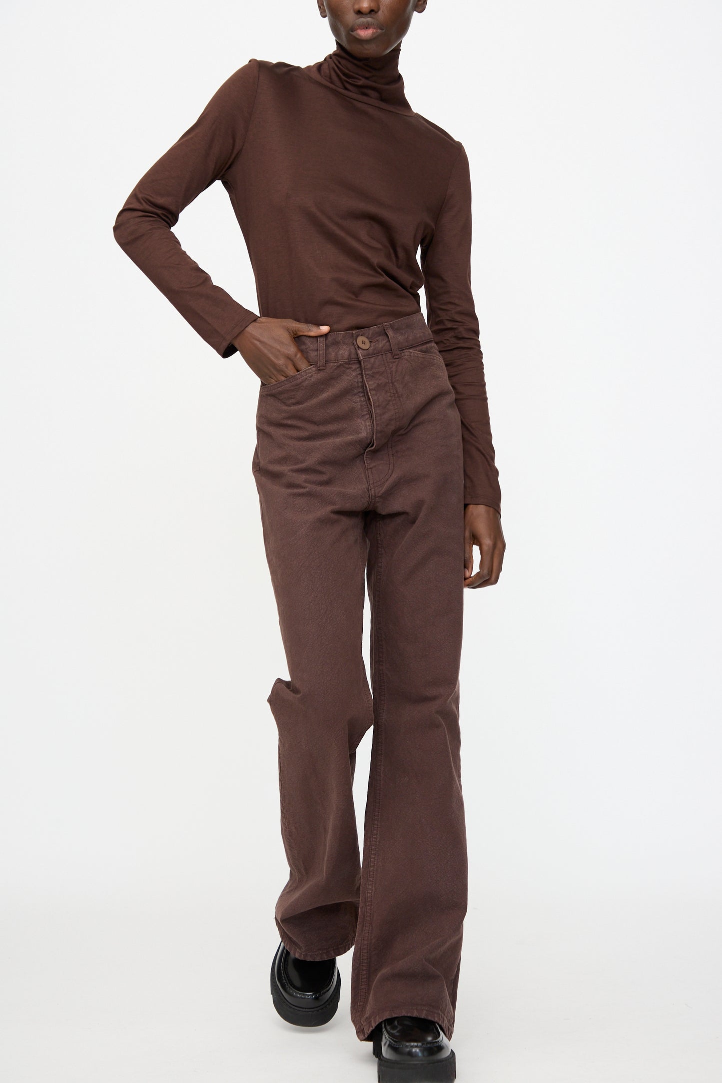 A person wearing Jesse Kamm’s Organic Canvas Les Deux Pants in Raisin with a brown turtleneck and black shoes stands against a plain white background.