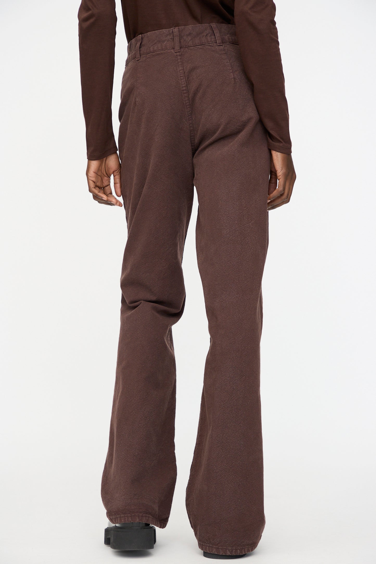 A person stands facing away from the camera, wearing Jesse Kamm’s Organic Canvas Les Deux Pants in Raisin and a long-sleeve brown top against a plain background.