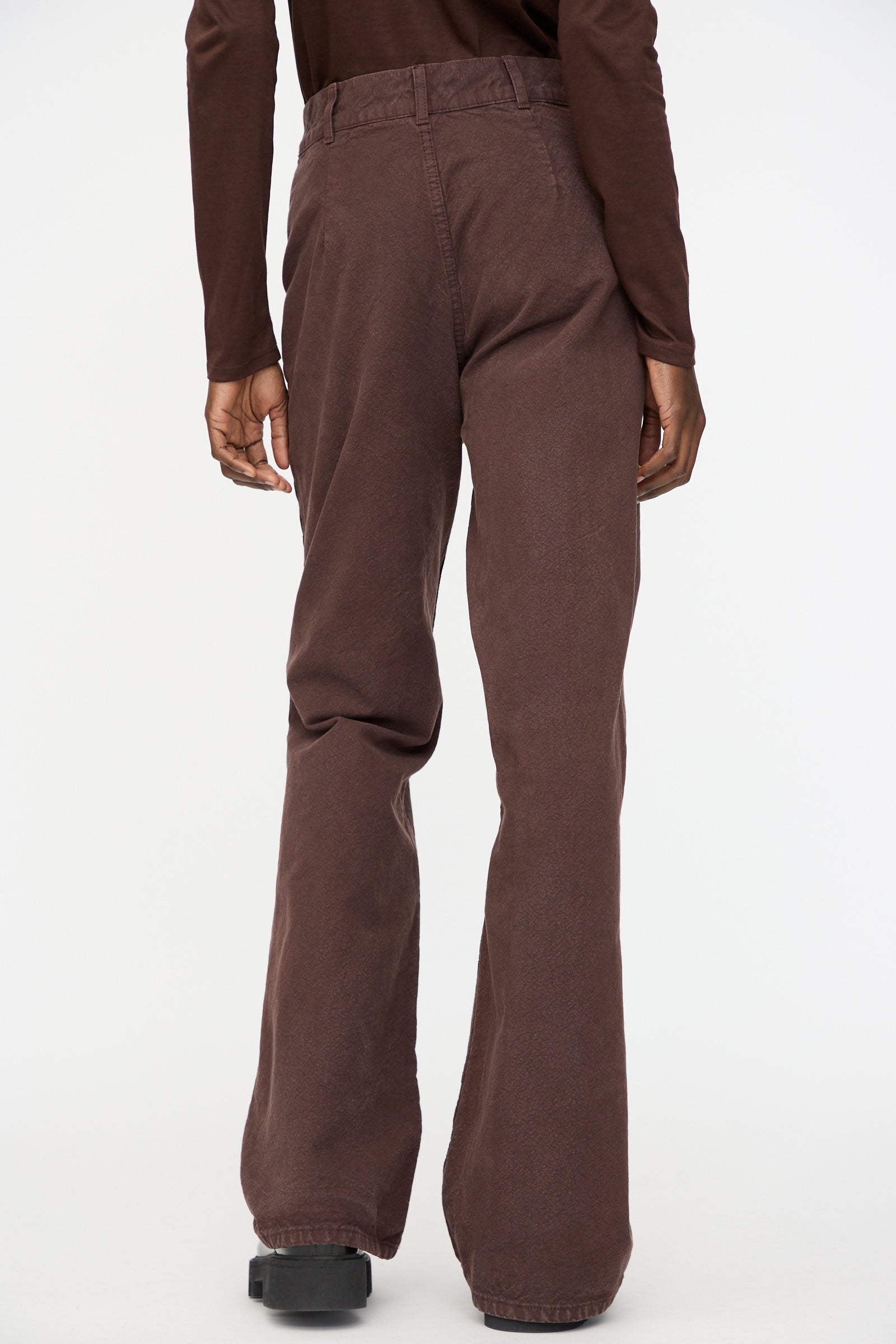 A person stands facing away from the camera, wearing Jesse Kamm’s Organic Canvas Les Deux Pants in Raisin and a long-sleeve brown top against a plain background.