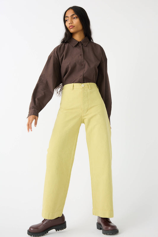 A person stands against a white background, wearing a brown long-sleeve shirt, Jesse Kamm's Organic Canvas Sailor Pant in Citroen, and brown boots. They have long dark hair and a neutral expression.