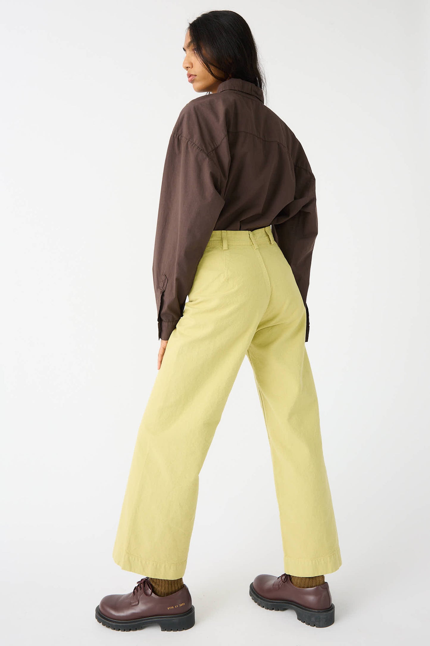 A person stands with their back to the camera, dressed in a brown shirt and Jesse Kamm's Organic Canvas Sailor Pant in Citroen, complemented by brown shoes, all against a plain white background.