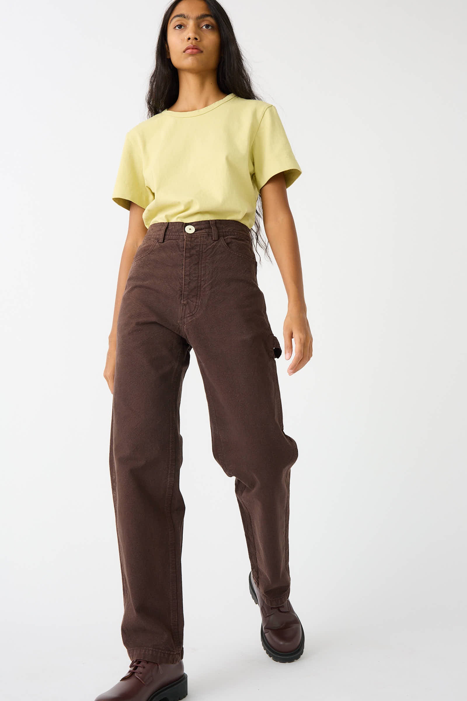 A person wearing a yellow t-shirt, the Organic Cotton Canvas Handy Pant in Raisin by Jesse Kamm, and dark brown shoes is standing against a plain white background.