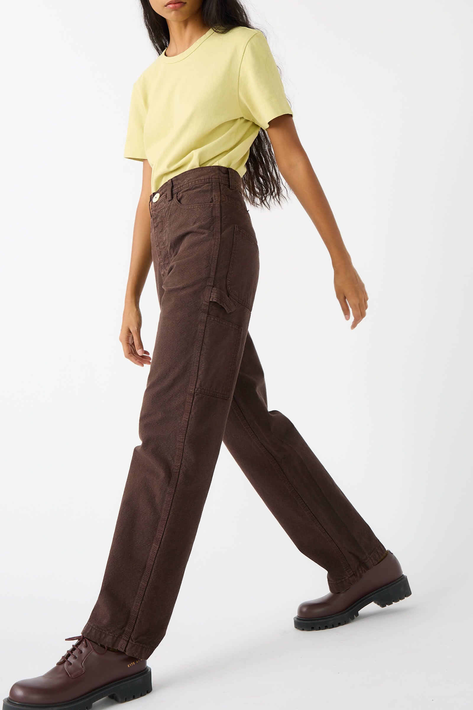 Person wearing a light yellow t-shirt, Jesse Kamm Organic Cotton Canvas Handy Pant in Raisin, and dark brown boots, walking with one foot slightly raised.