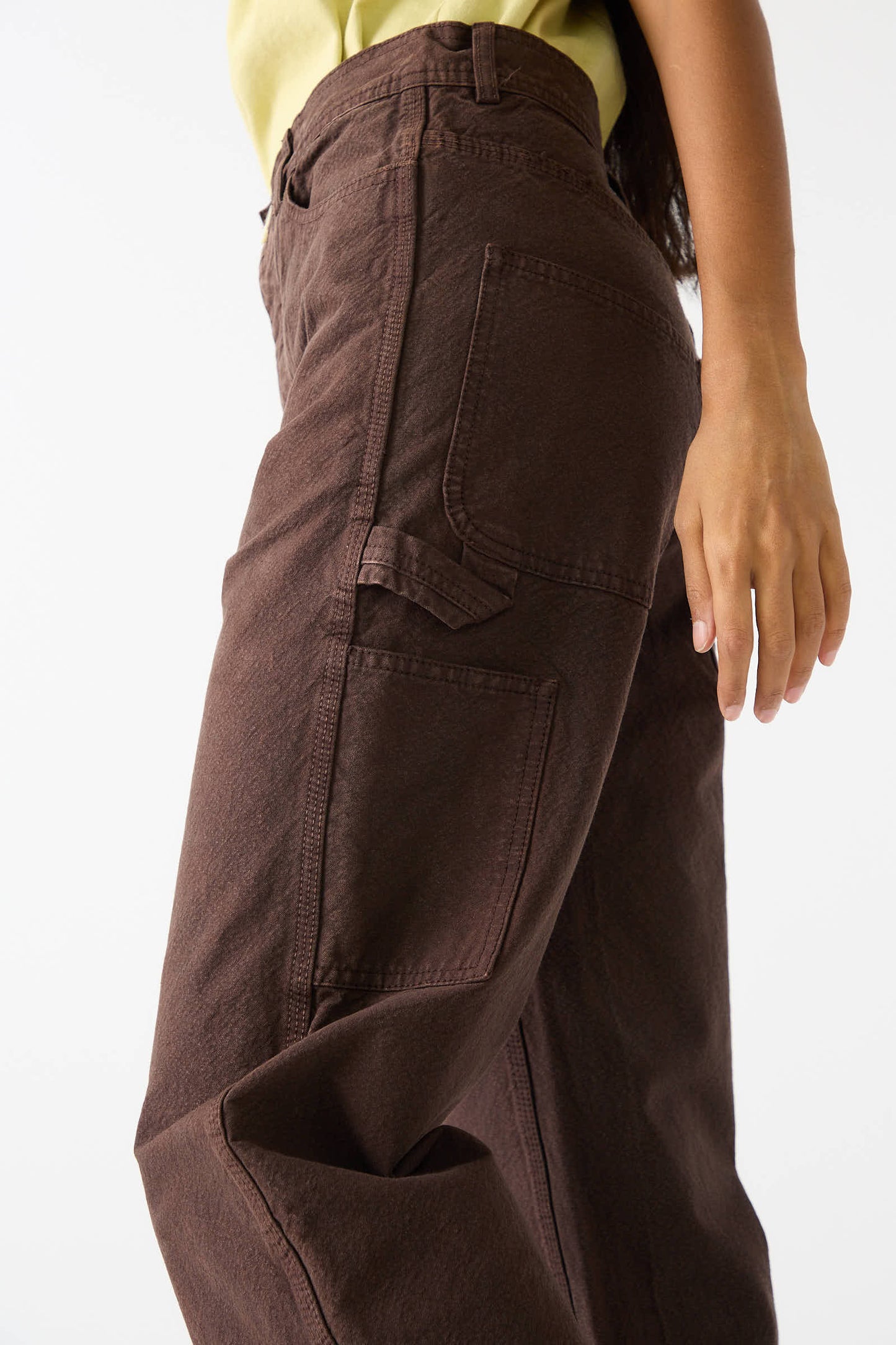 A person wearing Jesse Kamm's Organic Cotton Canvas Handy Pant in Raisin and a light yellow top is shown from the waist down with their hand resting on the side of their hip.