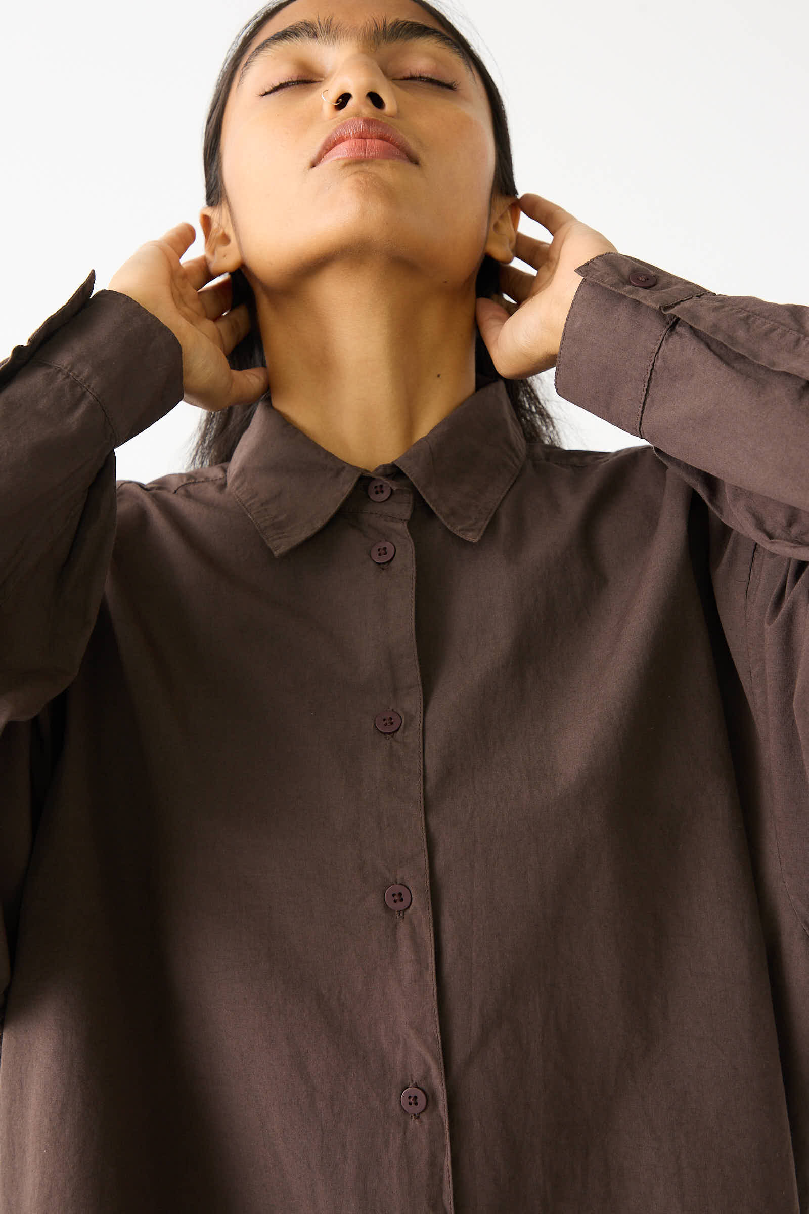 A person wearing a Jesse Kamm Organic Poplin Alberta Shirt in Raisin stands with eyes closed and head tilted back, hands resting on the back of their head.