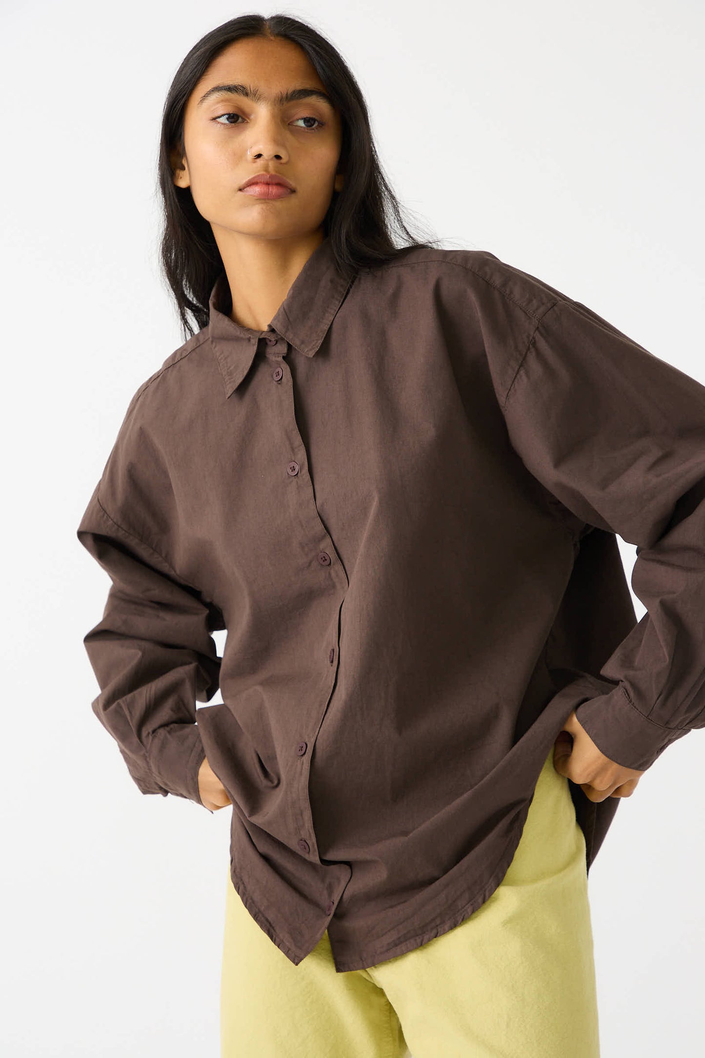 A person with long dark hair is wearing the Organic Poplin Alberta Shirt in Raisin by Jesse Kamm and light yellow pants, standing against a plain white background.