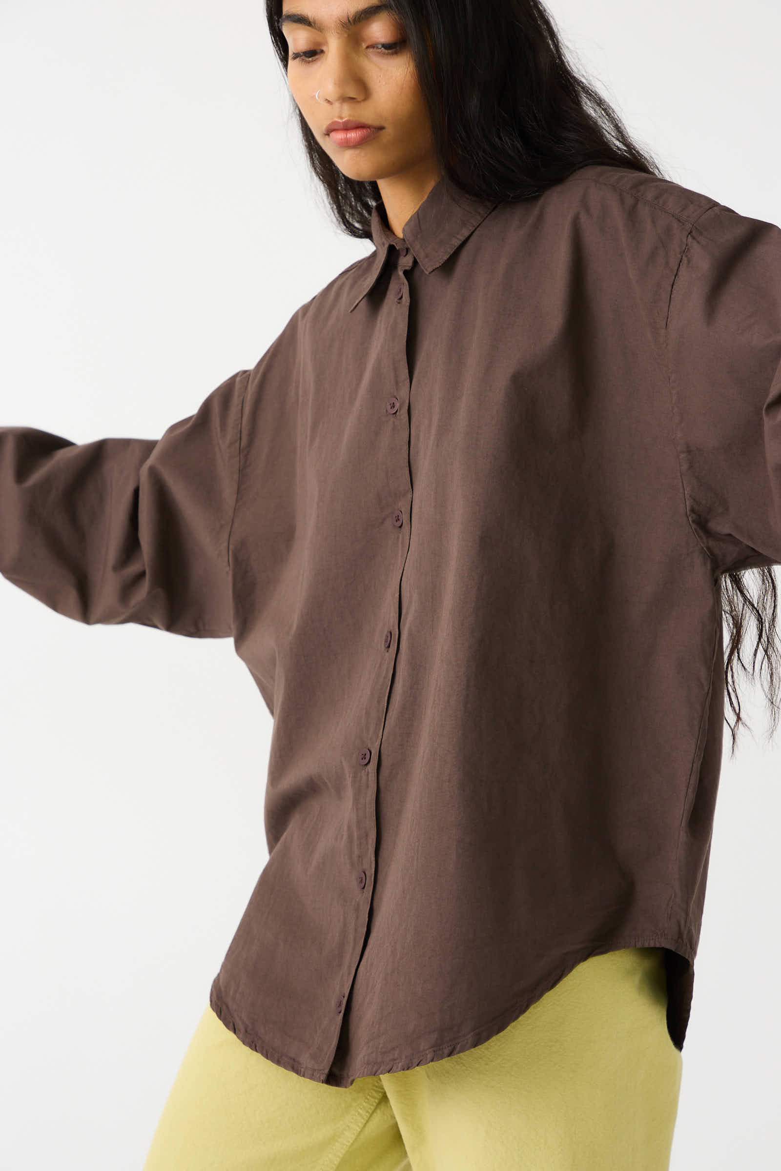 A person with long hair wears the loose-fitting Jesse Kamm Organic Poplin Alberta Shirt in a warm Raisin hue paired with light green pants, standing against a plain background.