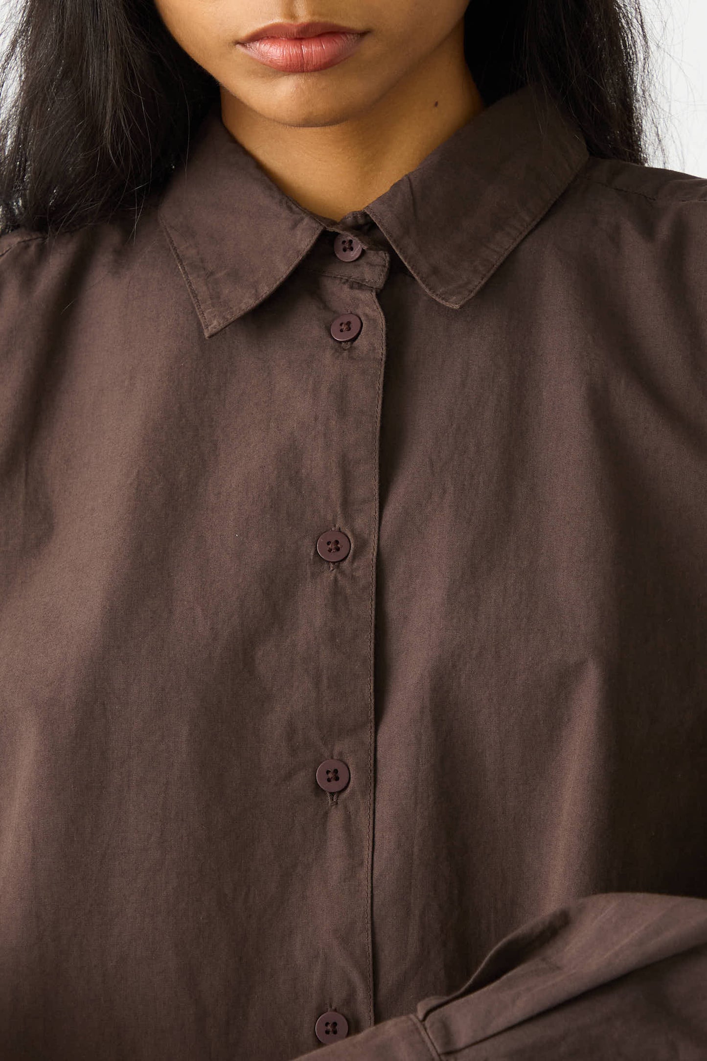 Close-up of a person wearing the Organic Poplin Alberta Shirt in Raisin by Jesse Kamm. The relaxed fit shirt, made from organic cotton, features a collar and six visible buttons, with the person's face partially visible.