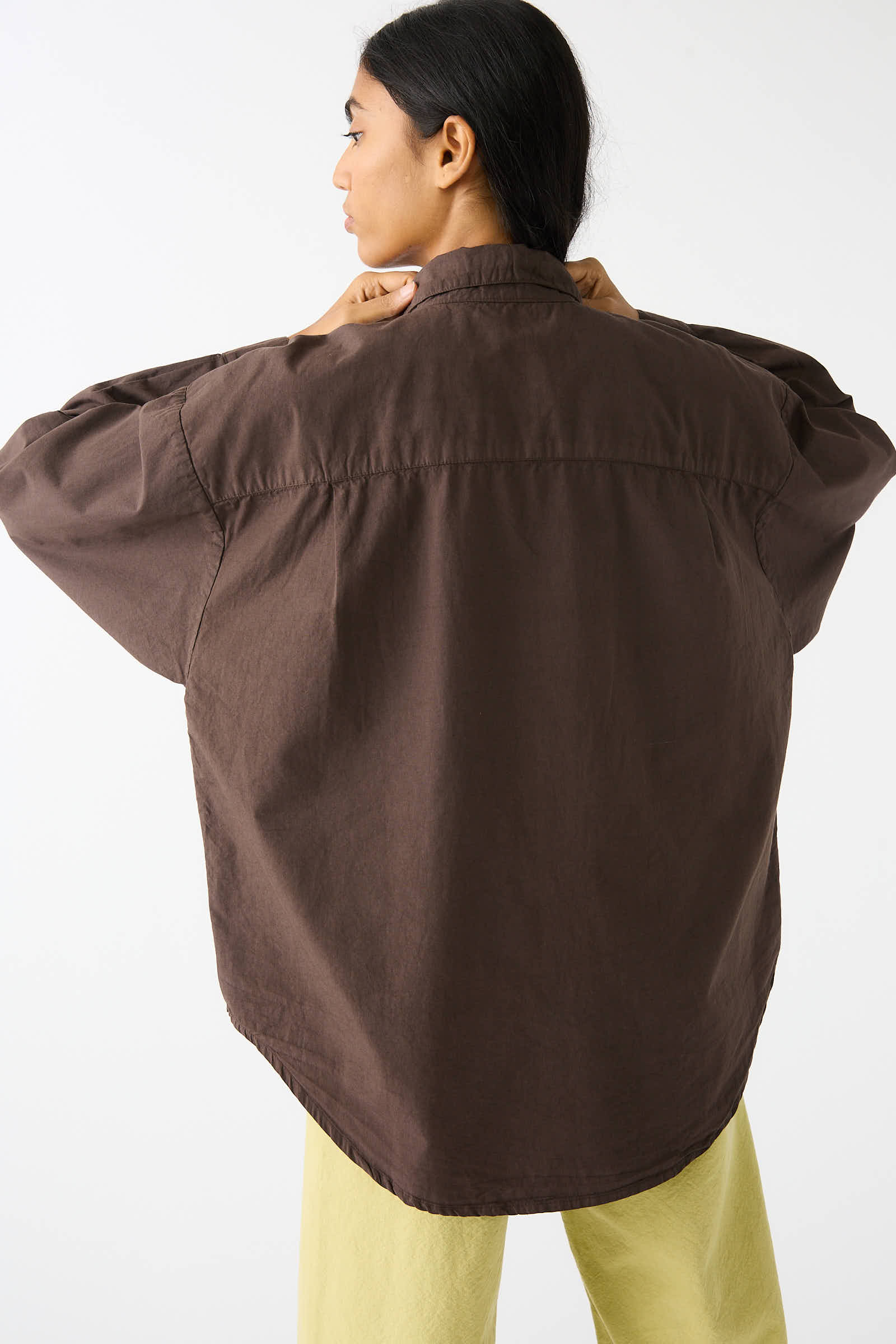 A person with long hair, seen from the back, adjusts the collar of a relaxed fit Organic Poplin Alberta Shirt in Raisin by Jesse Kamm. They are wearing light yellow pants and standing against a plain white background.