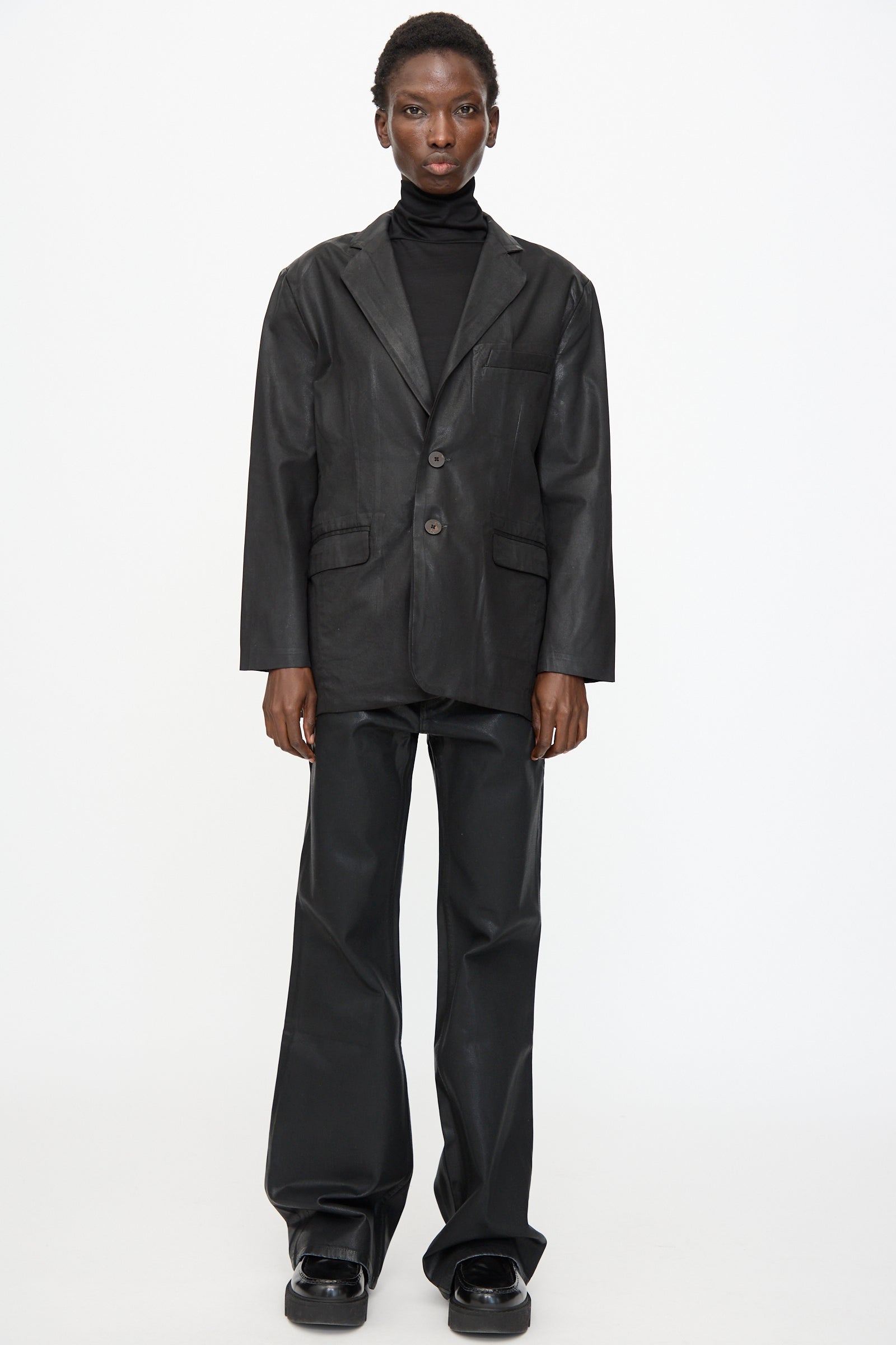 A person stands facing forward, wearing the Jesse Kamm Organic Twill Fianna Blazer in Black Lacquer, a black turtleneck, wide-leg black trousers, and black shoes against a plain white background.