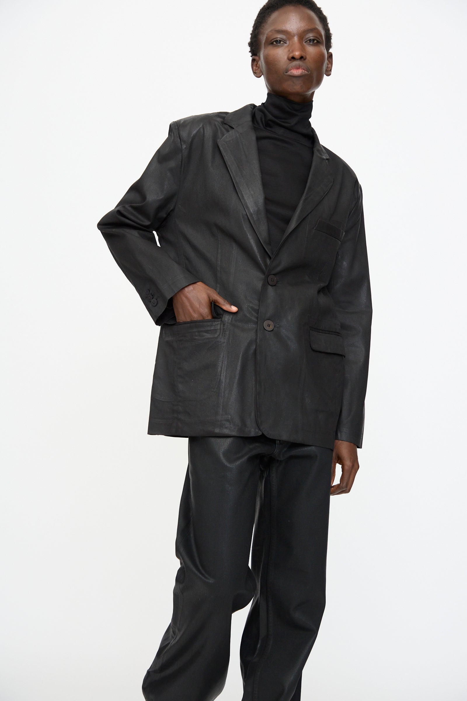 Model wears the Jesse Kamm Organic Twill Fianna Blazer in Black Lacquer over a black turtleneck and black pants, standing against a plain white background with one hand in their pocket.