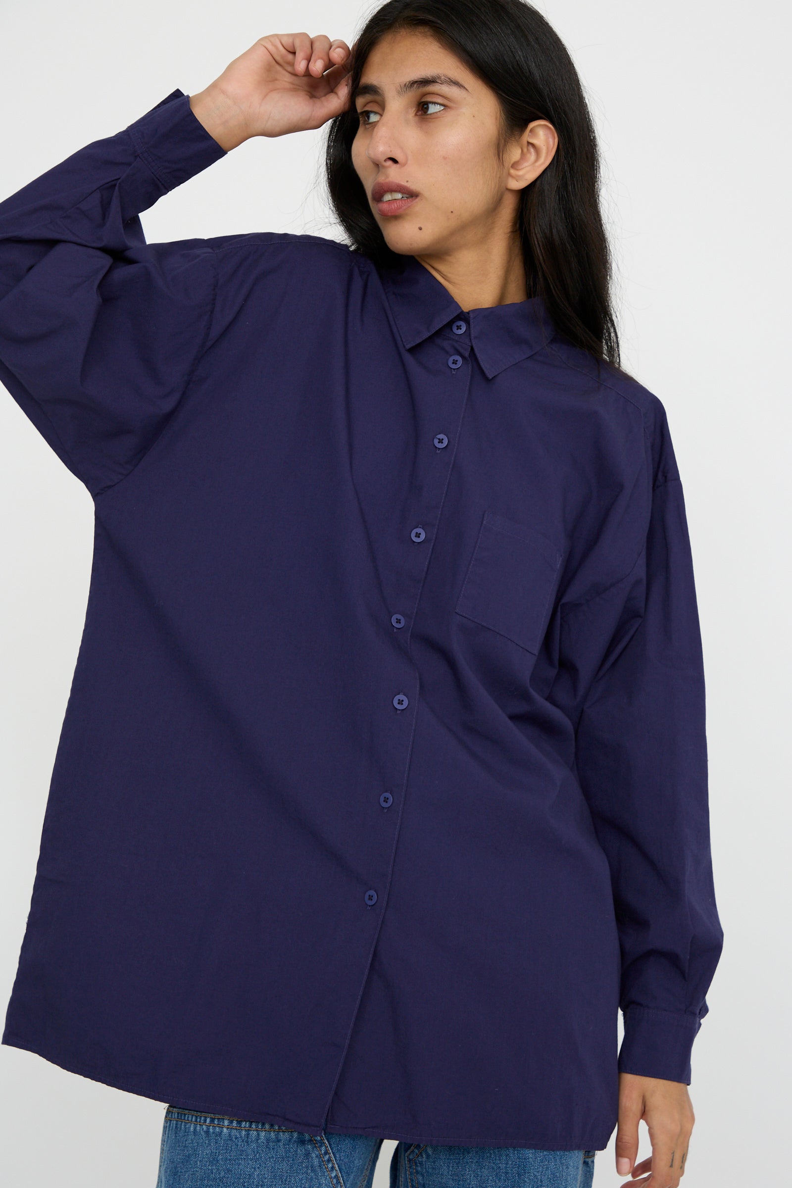 Against a plain background, a person is dressed in the oversized Painter Shirt in Bill Cunningham Blue by Jesse Kamm, made from soft cotton poplin, with one hand raised. 