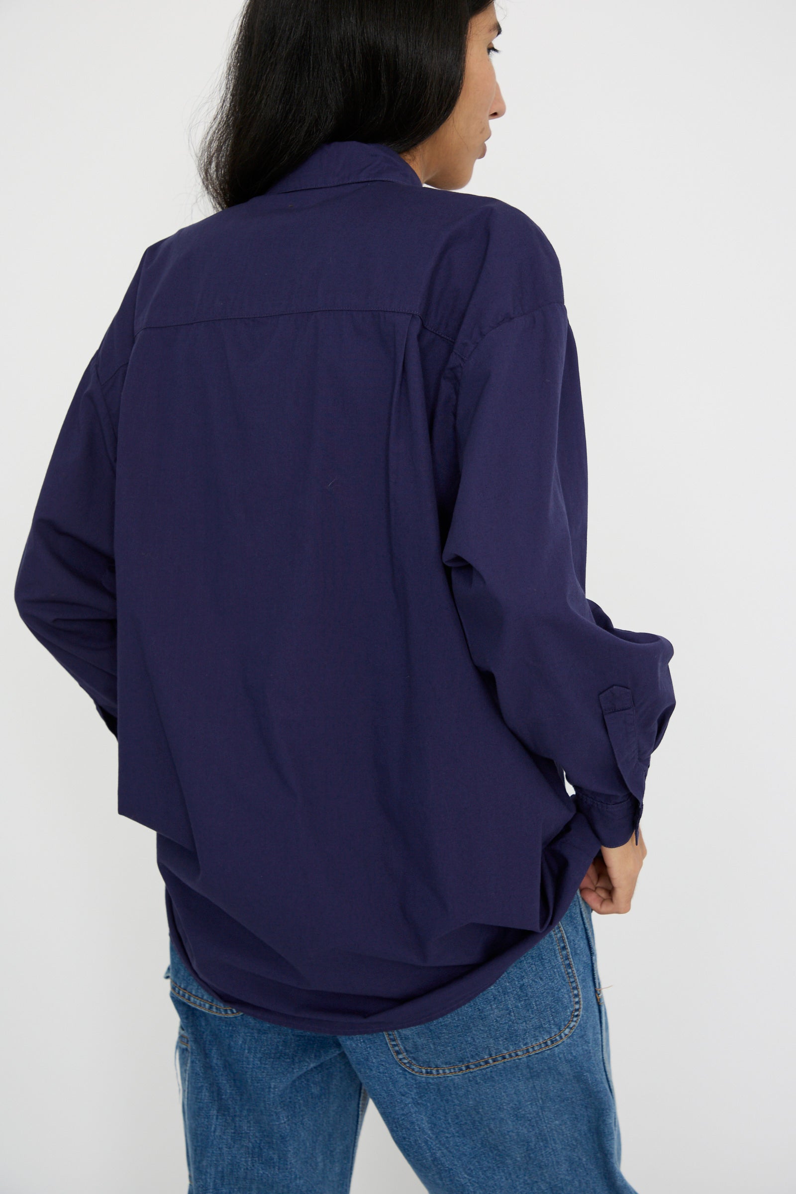 A person with long dark hair is wearing a loose-fitting Painter Shirt in Bill Cunningham Blue by Jesse Kamm and blue jeans, viewed from behind against a plain background.