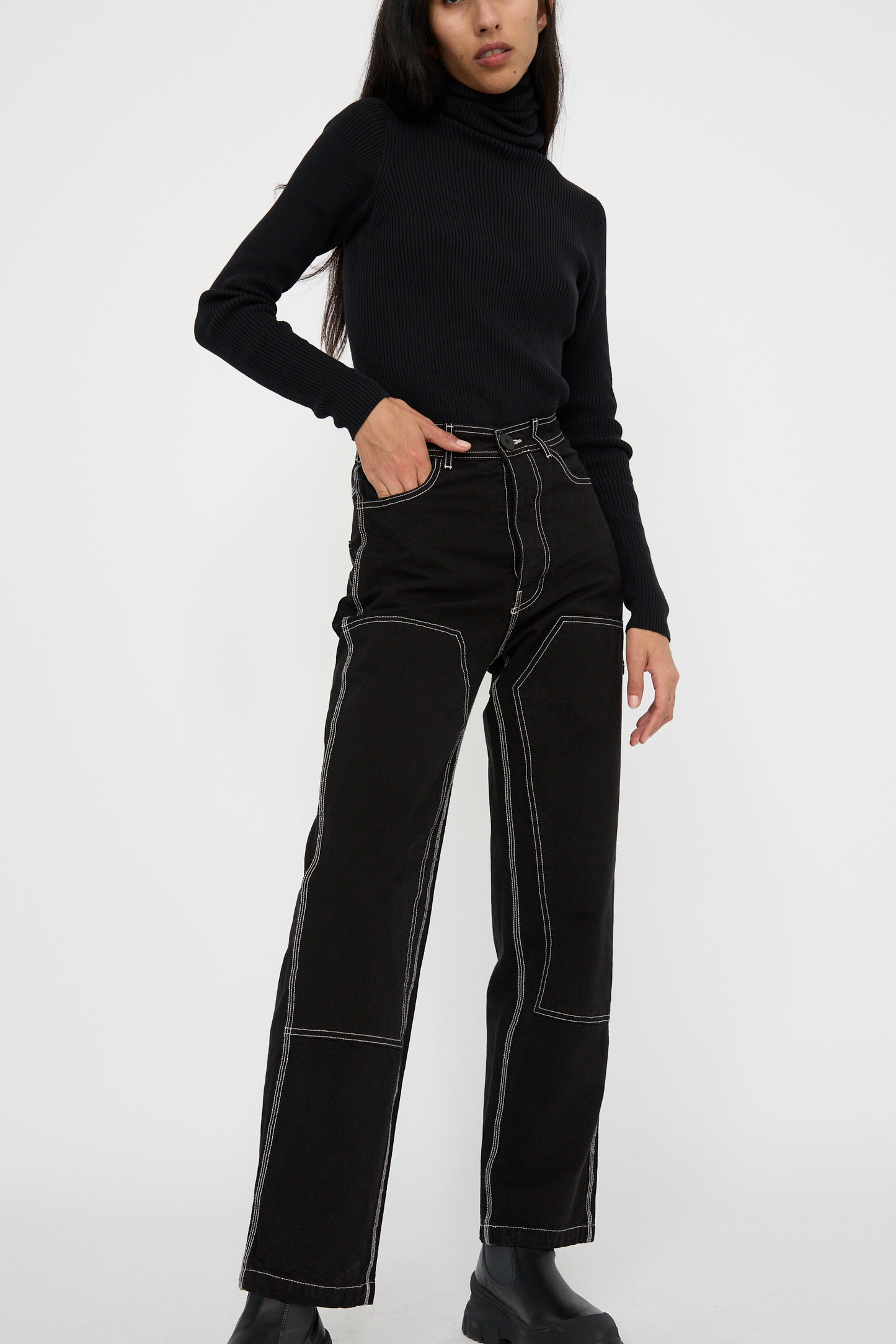 A person stands against a plain background, in a black turtleneck and Jesse Kamm's Patchfront Handy Pant in black with natural stitching. One hand rests in their pocket.