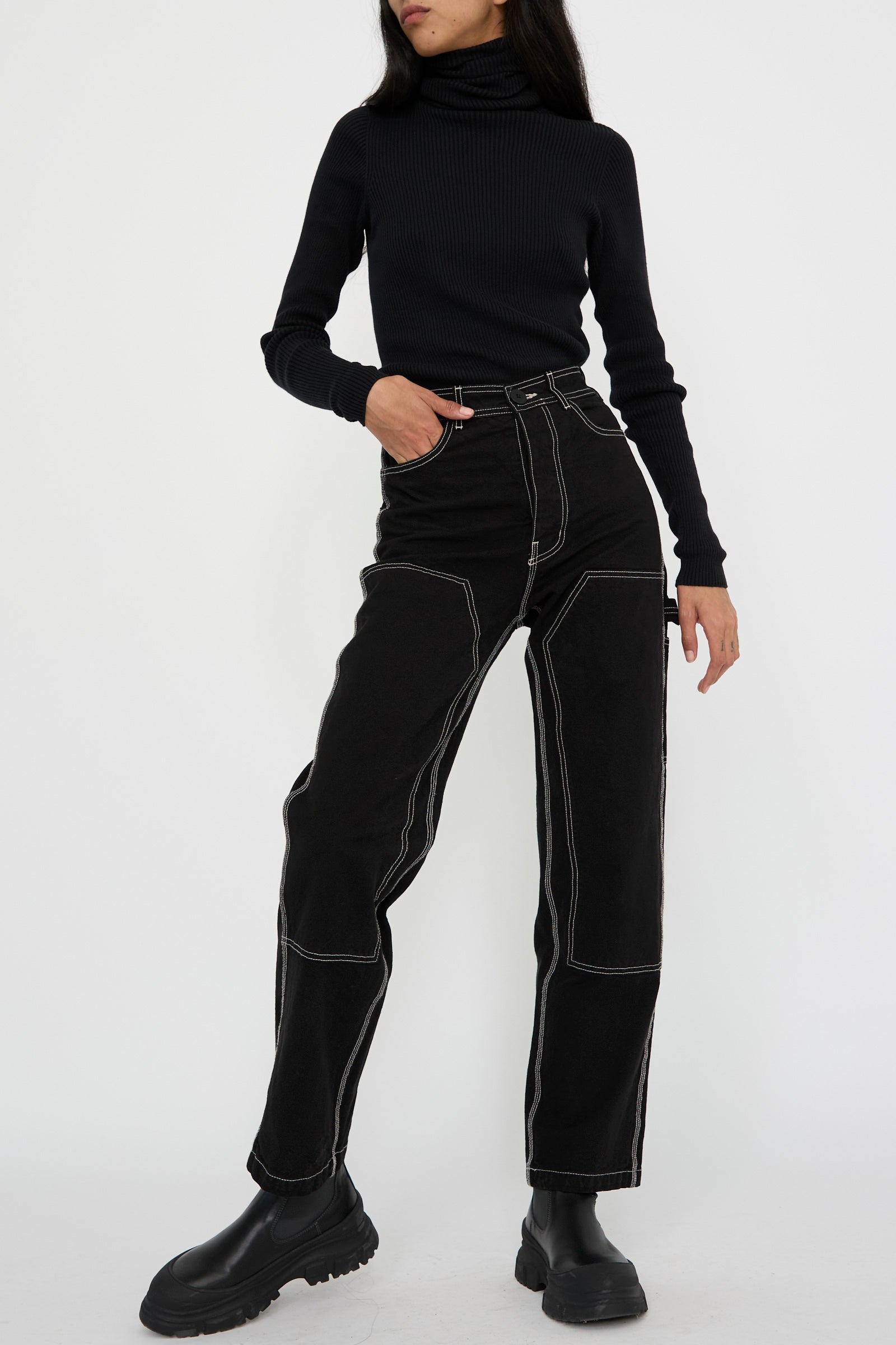 A person wearing a black turtleneck and the Patchfront Handy Pant in Black with Natural Stitching from Jesse Kamm, paired with sleek black boots, standing against a plain background with a hand in their pocket. 