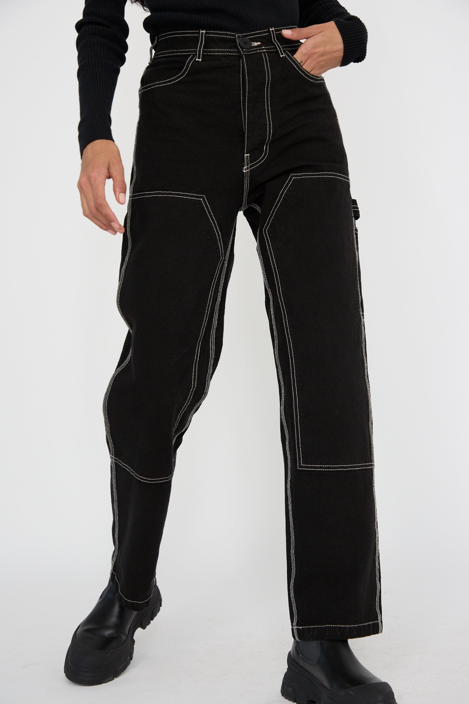 A person wearing the Jesse Kamm Patchfront Handy Pant in Black with Natural Stitching and black boots stands against a white background from the waist down.