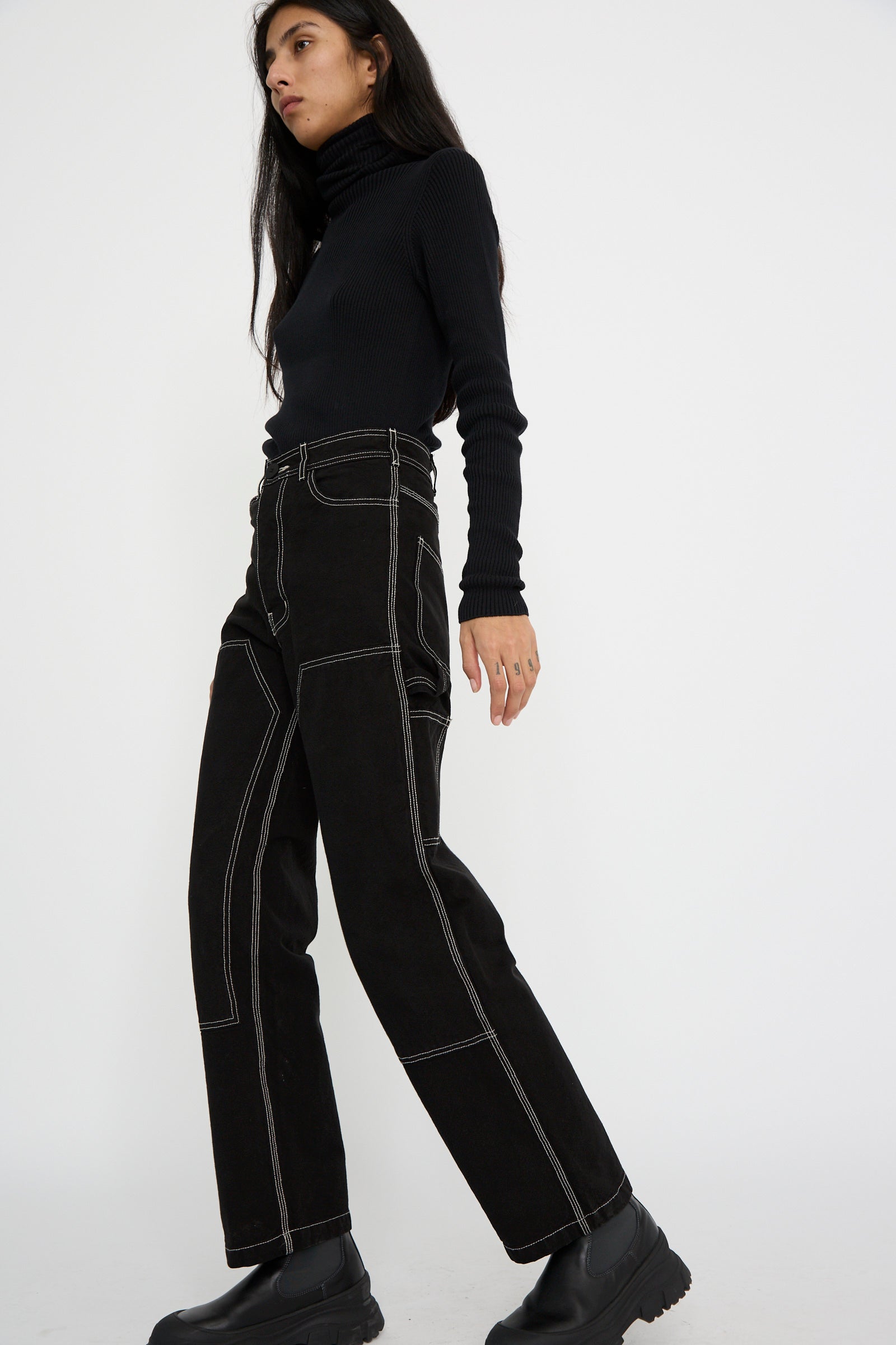 A person wearing the Jesse Kamm Patchfront Handy Pant in black with natural stitching and a black turtleneck walks forward against a plain white background.