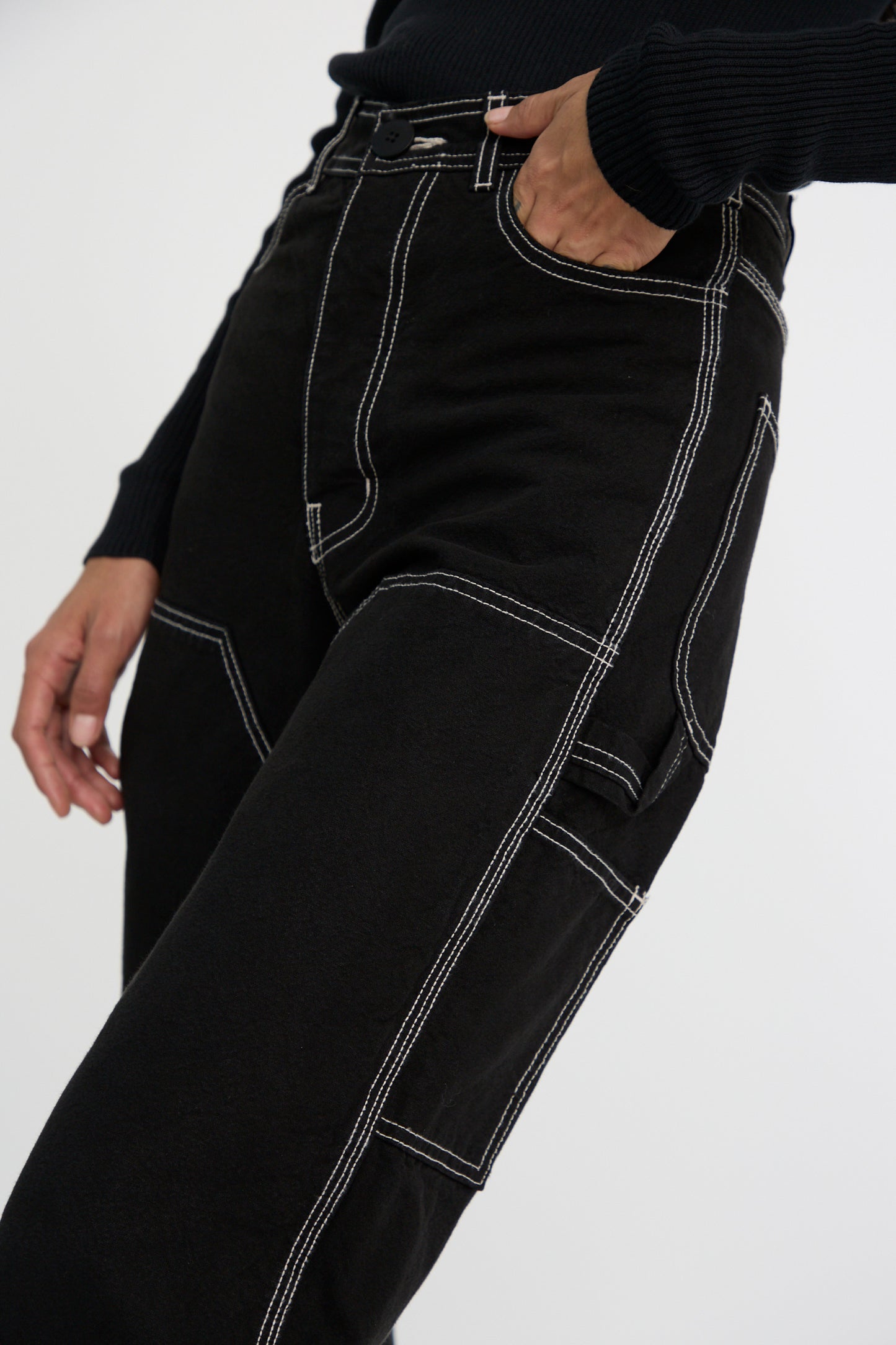 A person wears the Jesse Kamm Patchfront Handy Pant in Black with Natural Stitching, one hand tucked into a pocket, set against a plain background. 