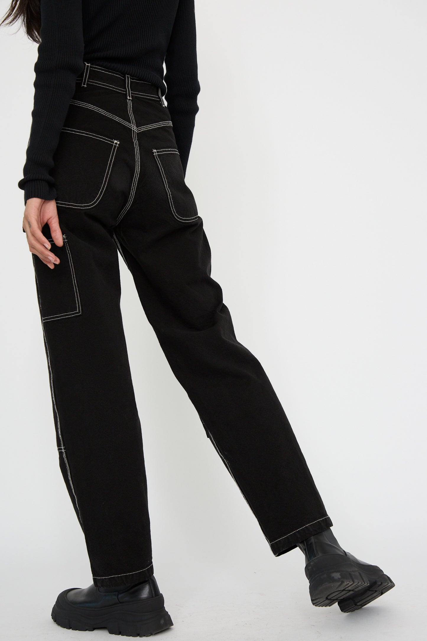 Sporting the Patchfront Handy Pant in Black with Natural Stitching by Jesse Kamm, a person wears them with a black long-sleeve top and sleek black boots, standing against a crisp white background.