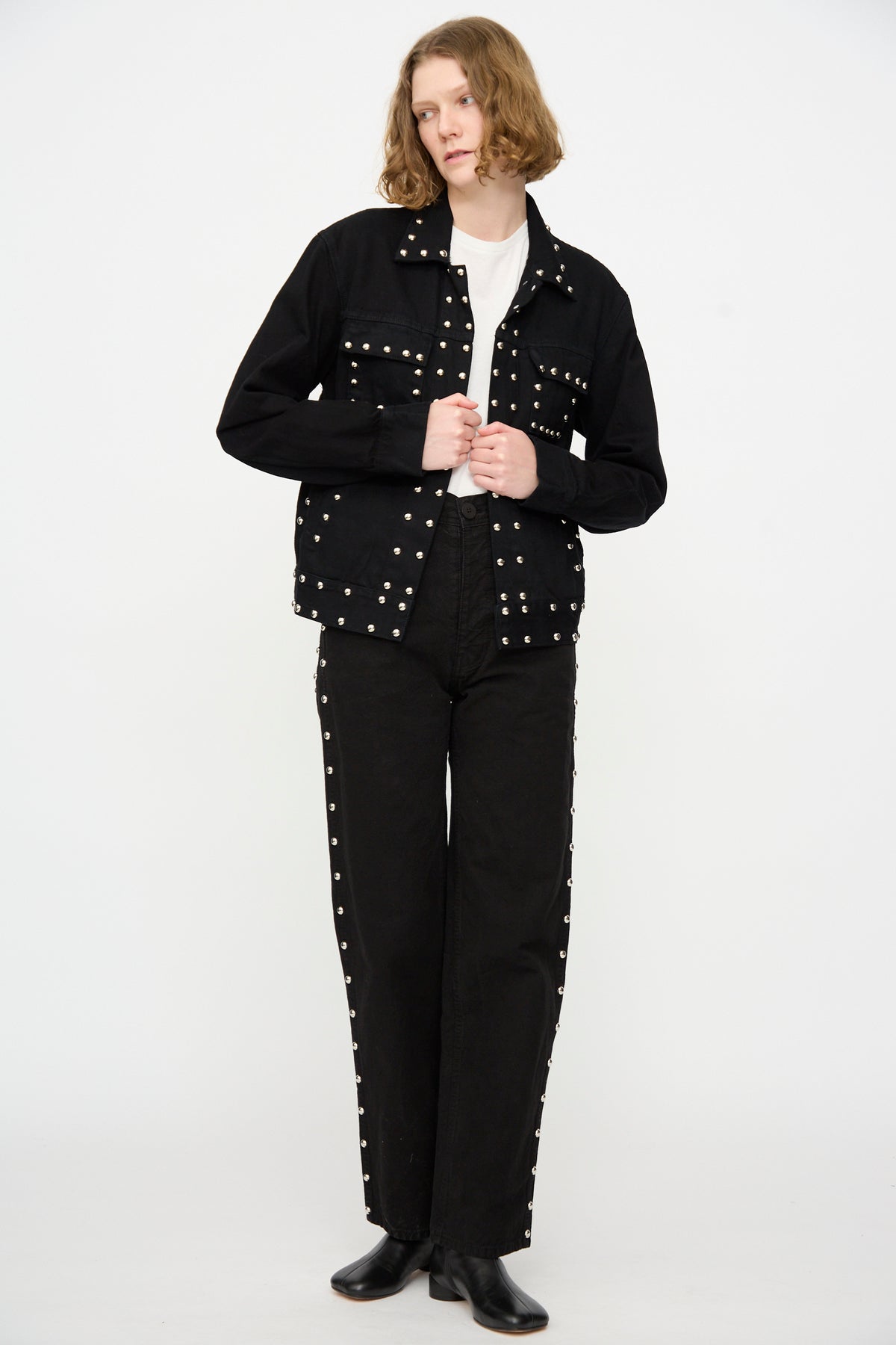 A person stands against a white background wearing the Jesse Kamm Ranch Jacket in Black Overdye with Studs, matching black studded pants, a white shirt, and black boots.