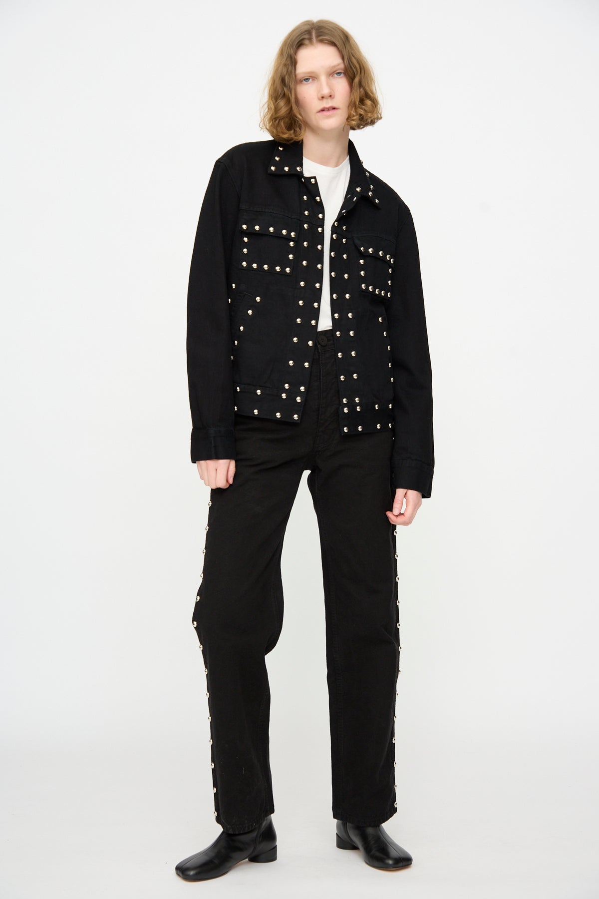 A person stands against a plain white background, wearing the Jesse Kamm Ranch Jacket in Black Overdye with Studs, paired with black pants, a white shirt, and black boots.