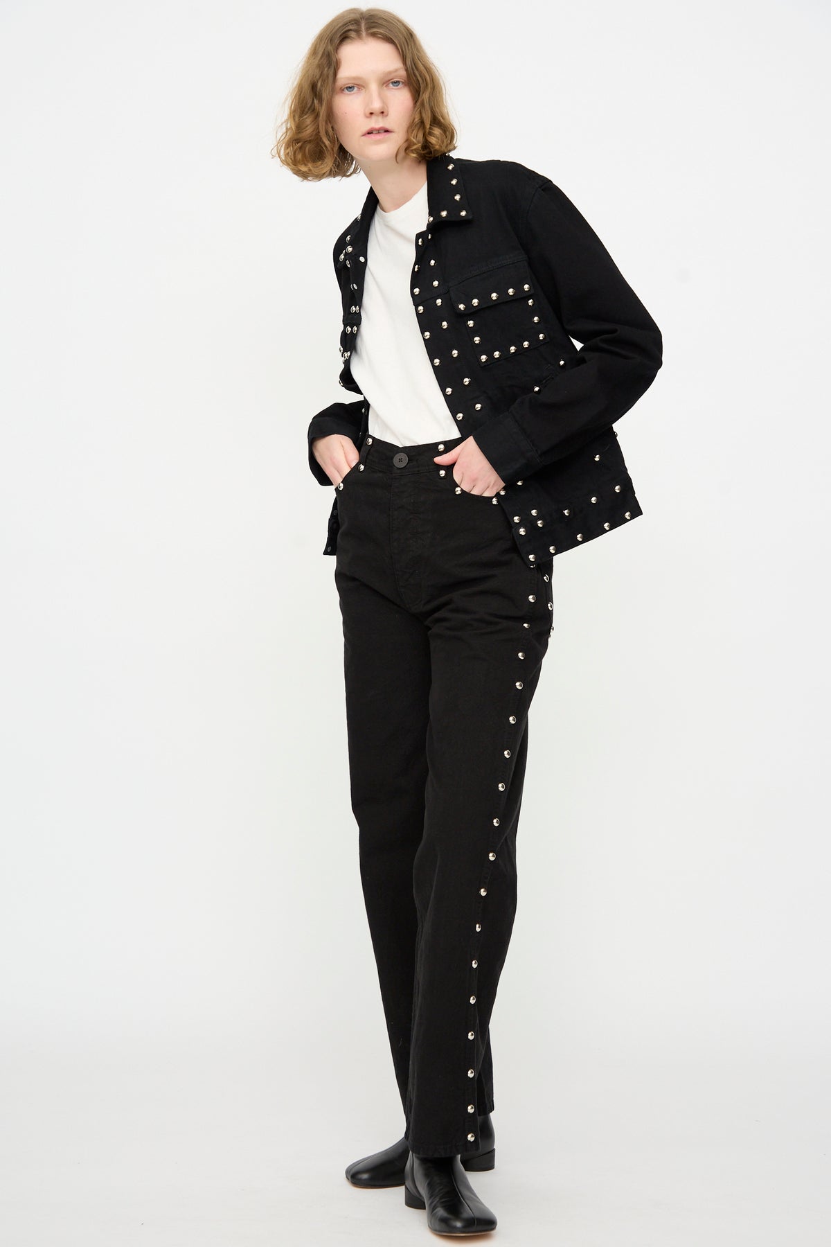 A person stands against a plain background wearing the Jesse Kamm Ranch Jacket in Black Overdye with Studs, paired with a white shirt, black studded pants, and black boots.