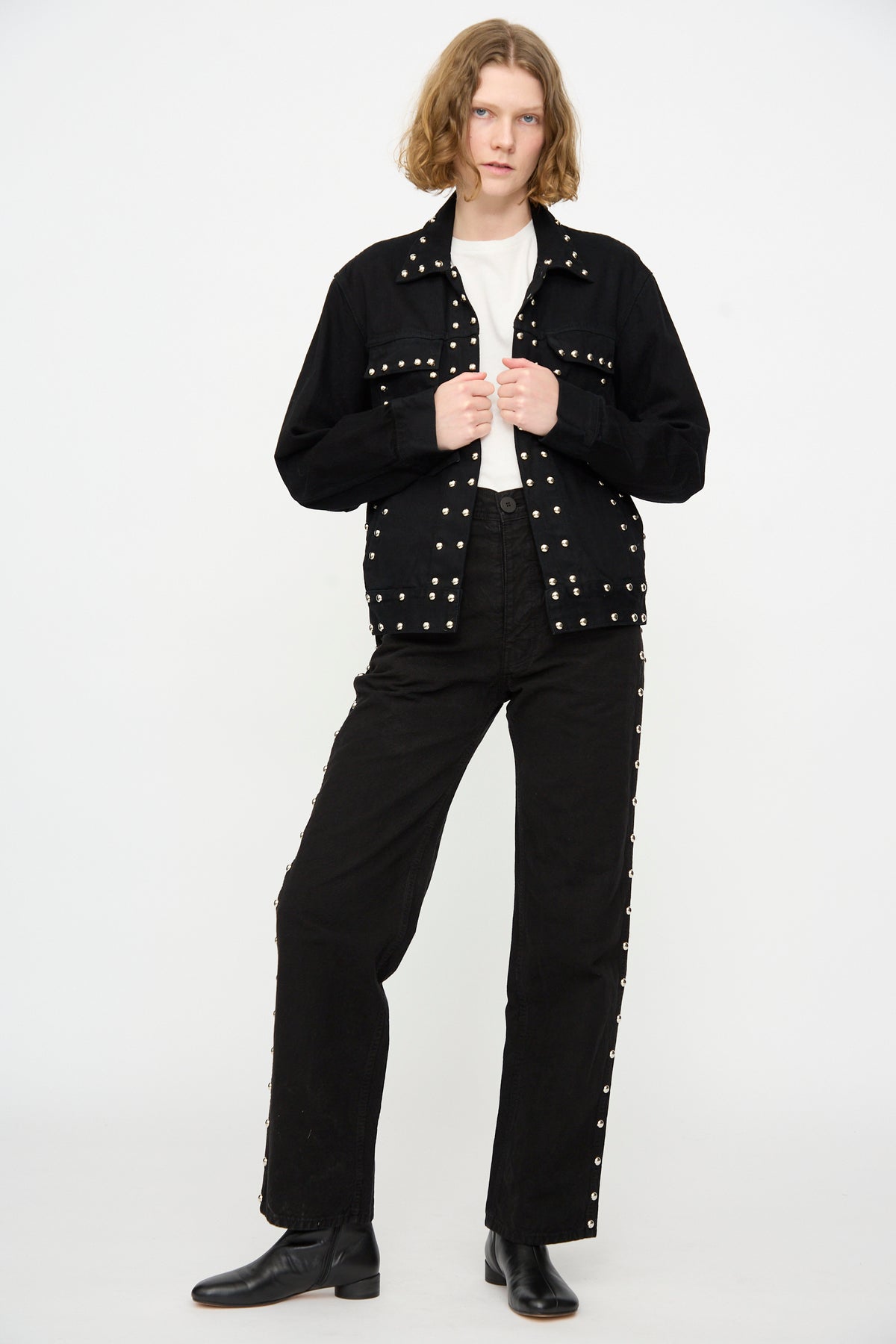 A person stands against a white background wearing the Jesse Kamm Ranch Jacket in Black Overdye with Studs, paired with black studded pants, a white shirt, and black boots.