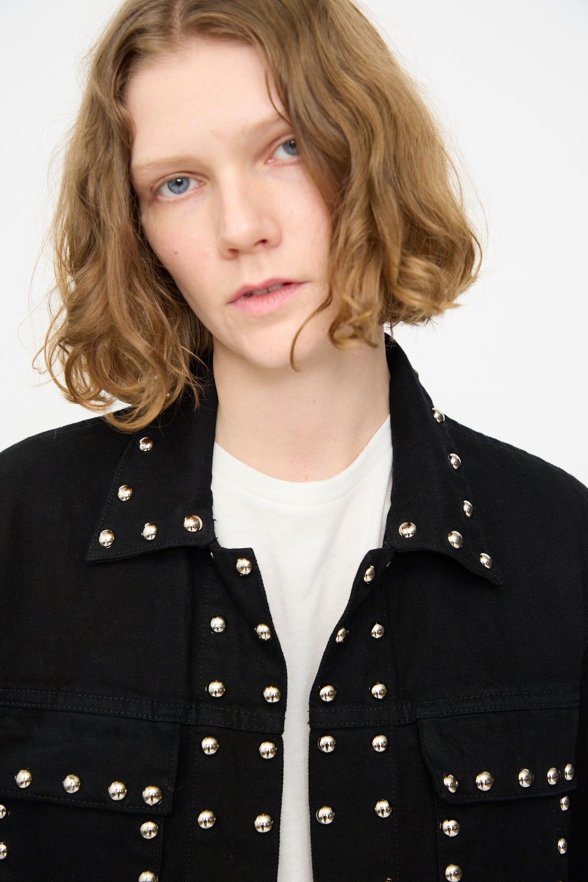 A person with short, wavy, light brown hair wears the Jesse Kamm Ranch Jacket in Black Overdye with Studs over a white shirt, gazing slightly past the camera against a plain light background.