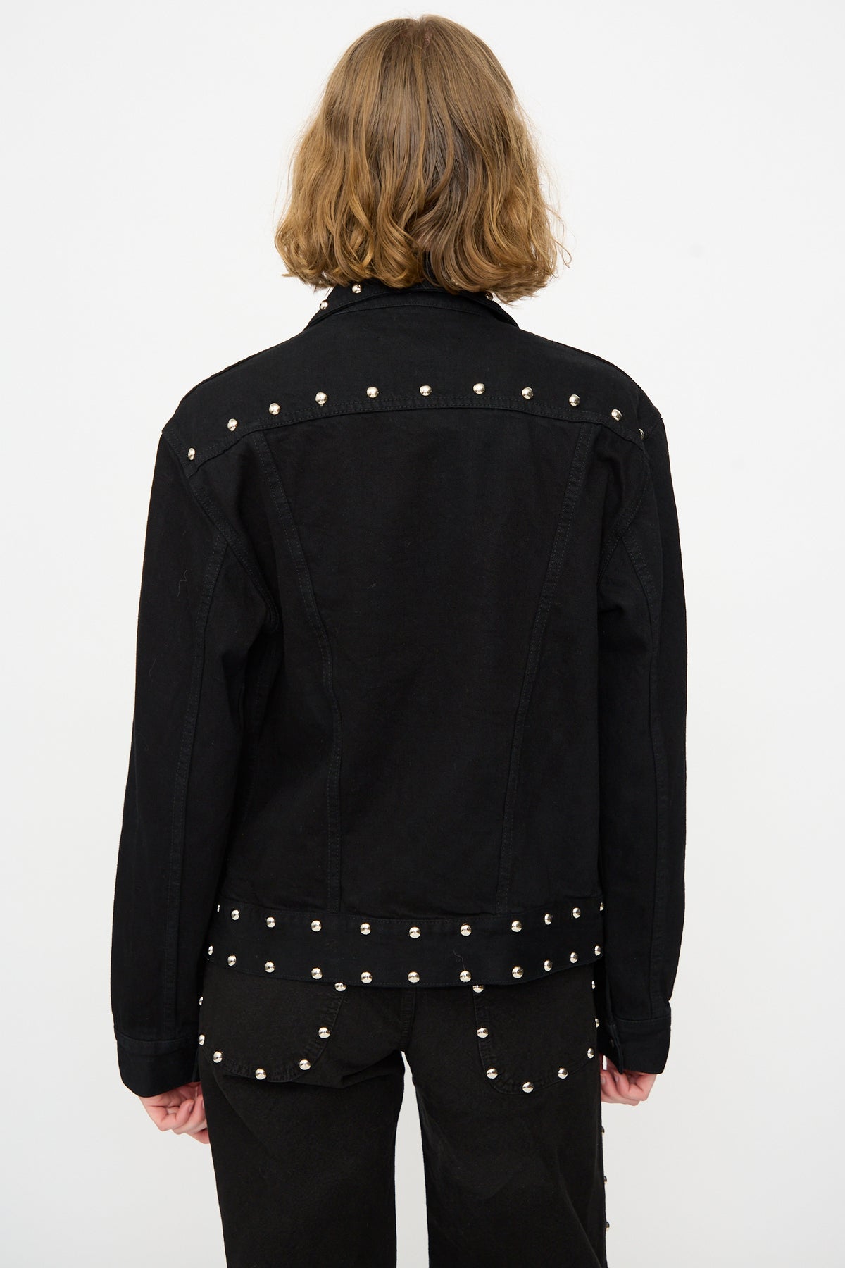 A person with shoulder-length light brown hair is seen from behind against a plain white background, wearing the Jesse Kamm Ranch Jacket in Black Overdye with Studs and matching studded pants.