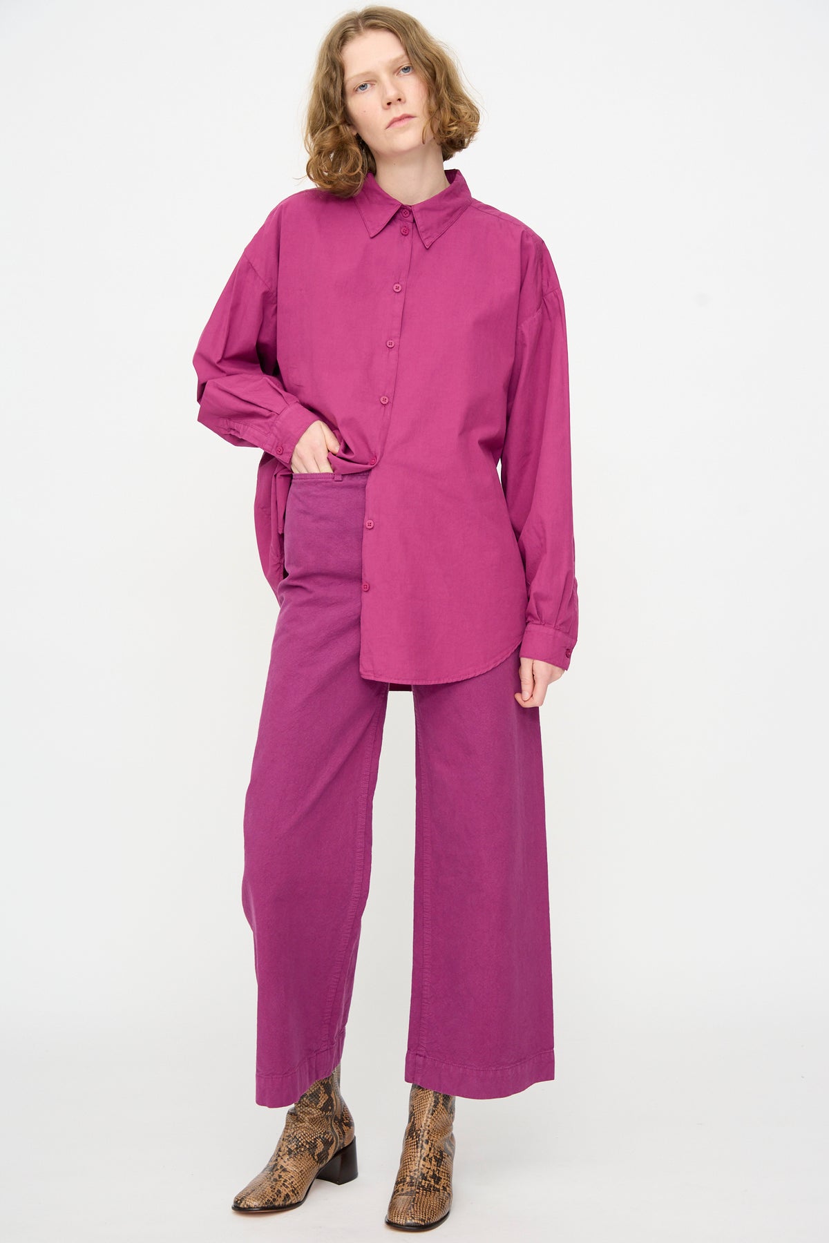 A person stands against a plain background, wearing a loose-fitting magenta button-up shirt paired with Jesse Kamm’s Sailor Pant in Beet, a high-waist style crafted from organic cotton, with one hand slipped into the side pocket. The look is finished with brown snakeskin boots.