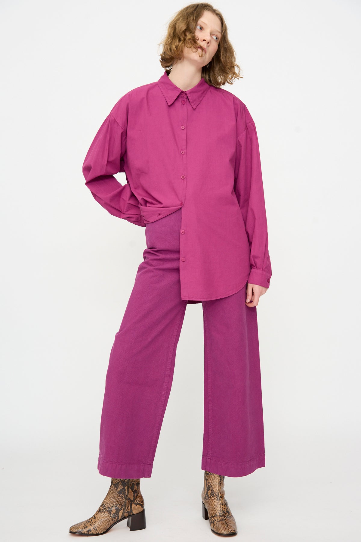 A person wearing a loose-fitting magenta shirt and Jesse Kamm’s high-waist, wide-leg Sailor Pant in Beet, made from organic cotton, paired with brown snakeskin ankle boots, standing against a plain white background.