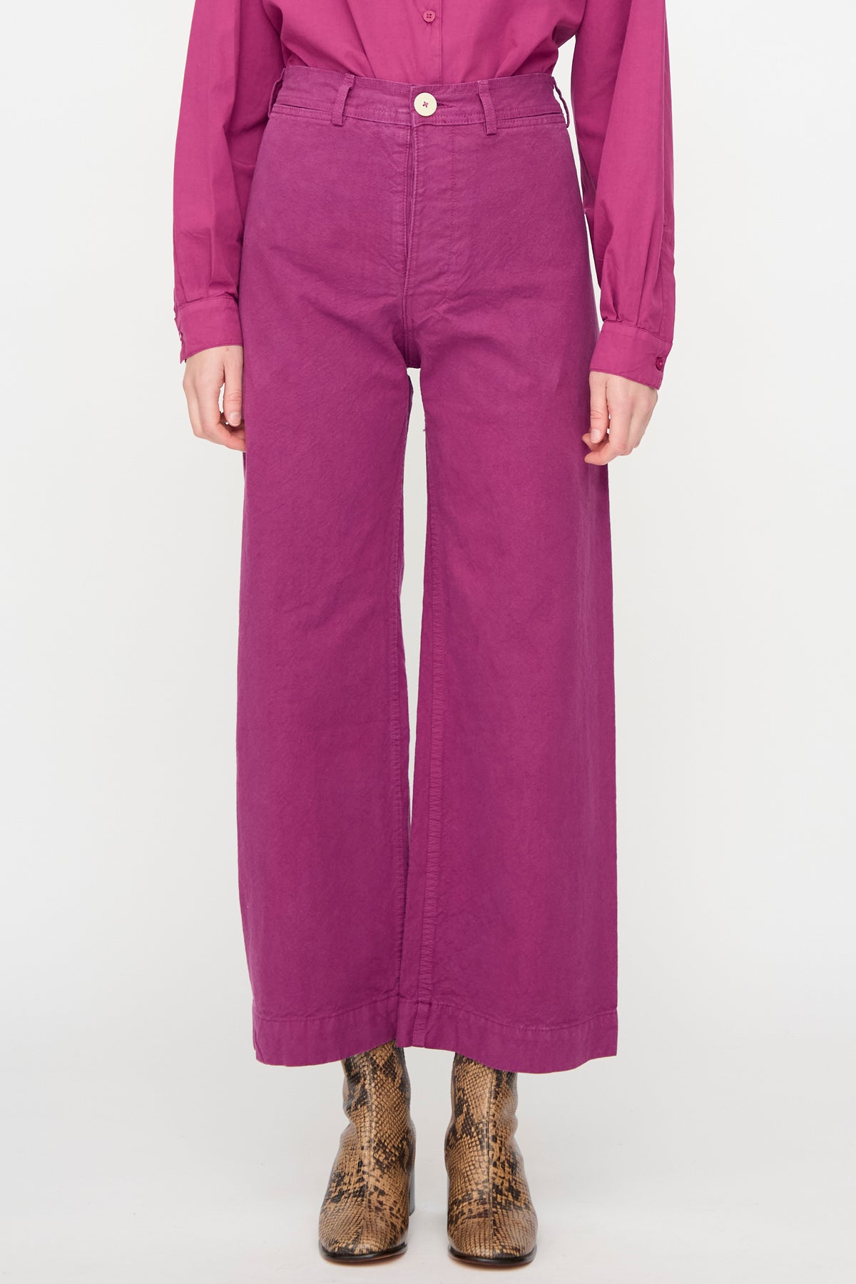 A person wearing a magenta button-up shirt and the Jesse Kamm Sailor Pant in Beet, crafted from organic cotton, paired with brown snake-patterned boots, stands against a plain white background.