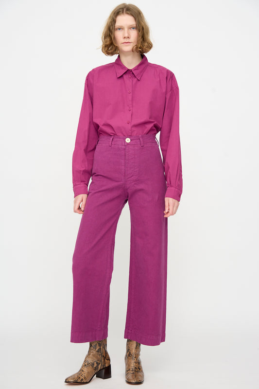 A person stands against a white background wearing a magenta button-up shirt, brown patterned ankle boots, and the Jesse Kamm Sailor Pant in Beet—an organic cotton, high-waisted style with wide legs.