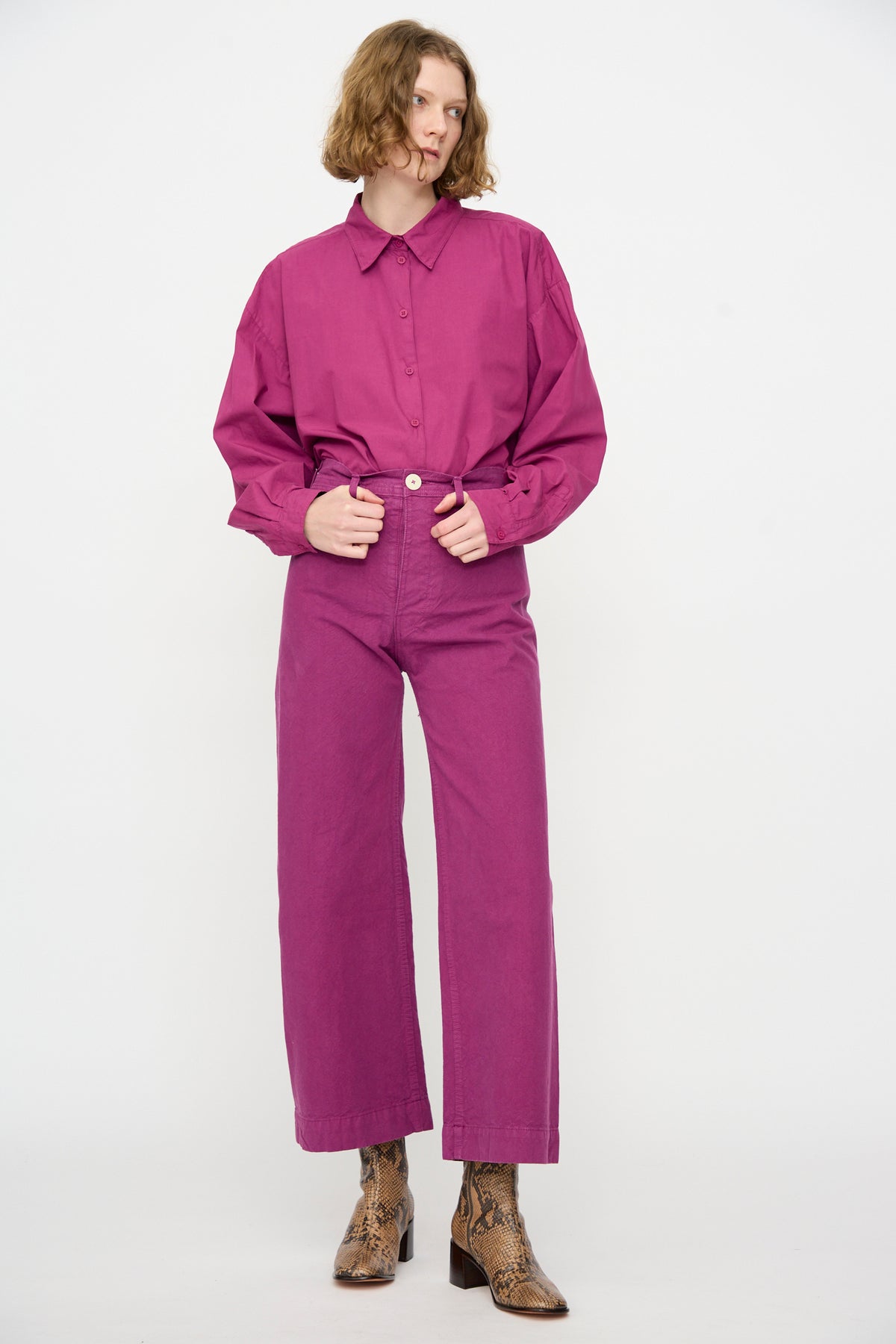 A person wearing a long-sleeved magenta shirt and the Jesse Kamm Sailor Pant in Beet stands facing forward, hands in pockets, paired with brown ankle boots.
