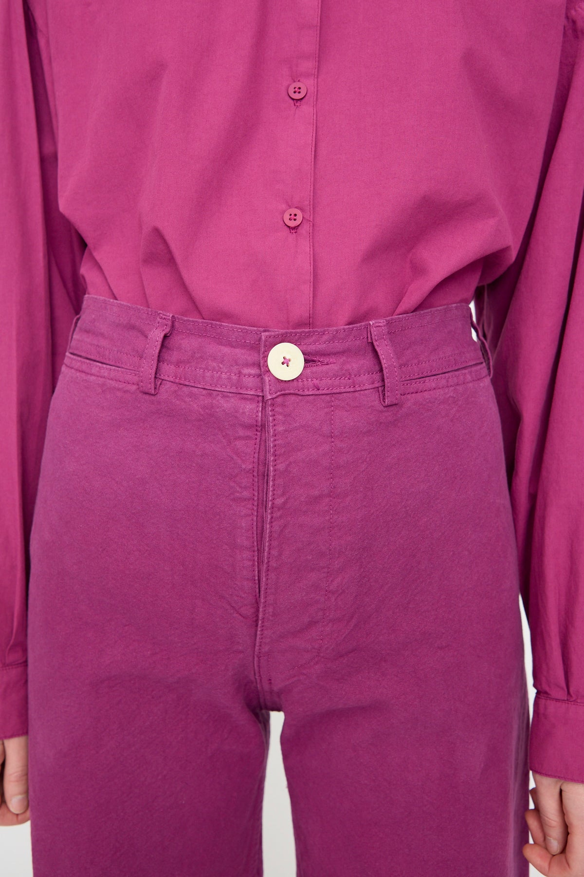 Person wearing a magenta button-up shirt tucked into Jesse Kamm’s Sailor Pant in Beet, crafted from organic cotton and finished with a light-colored button.