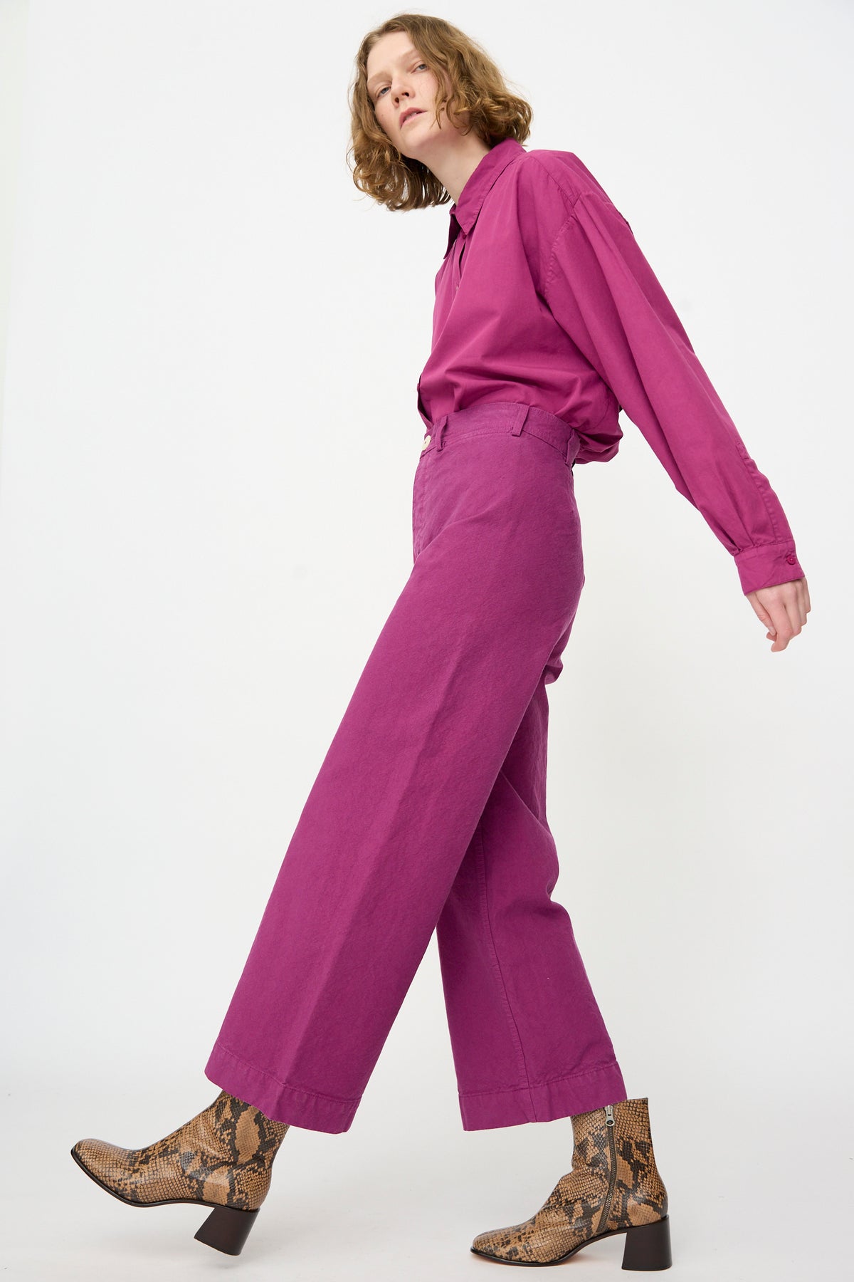 Wearing the Jesse Kamm Sailor Pant in Beet, paired with a matching magenta organic cotton button-up shirt and brown snakeskin ankle boots, the person poses against a plain white background.