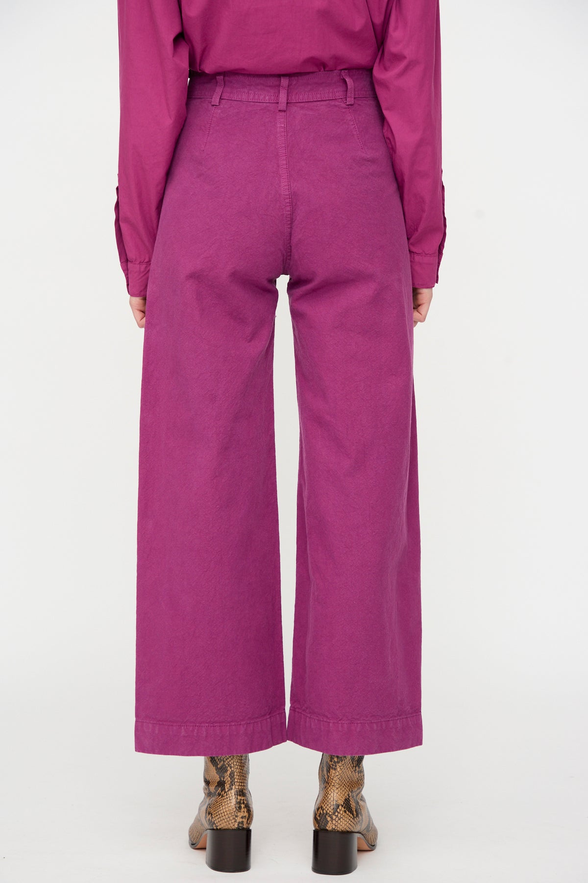 A person is shown from the back against a plain white background, wearing the Jesse Kamm Sailor Pant in Beet—a high waist, magenta wide-leg pant made from organic cotton—paired with a matching magenta shirt and snakeskin heeled boots.