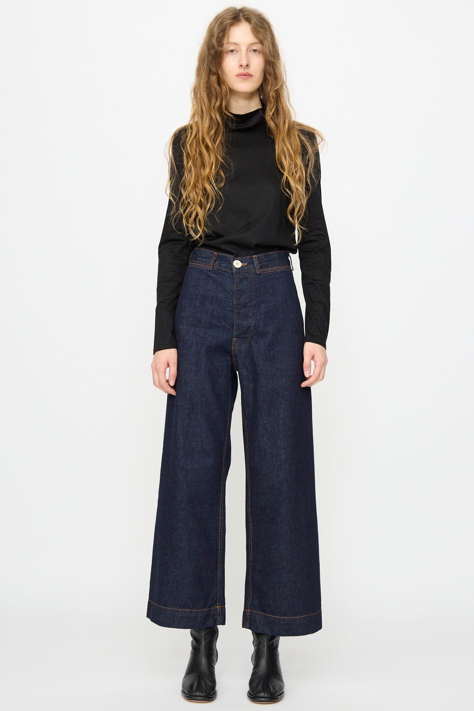 A person with long wavy hair wears a black long-sleeve top, Jesse Kamm Sailor Pant in Dark Blue, and black ankle boots, standing facing forward against a plain white background.