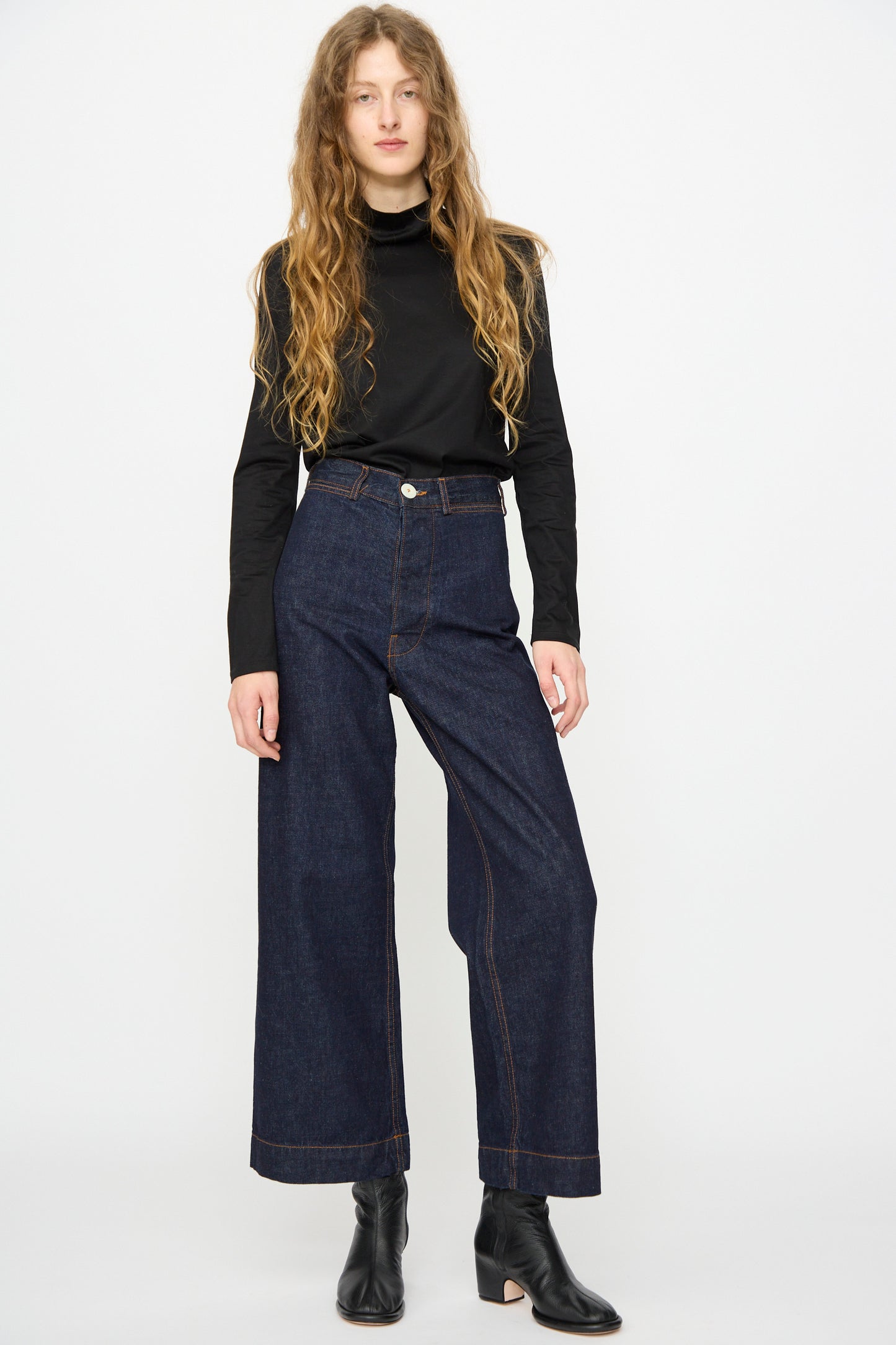 A person stands against a plain background wearing a black long-sleeve top, Jesse Kamm Sailor Pant in Dark Blue, and black ankle boots.