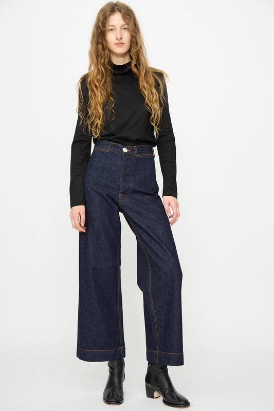 A person stands against a plain background wearing a black long-sleeve top, Jesse Kamm Sailor Pant in Dark Blue, and black ankle boots.
