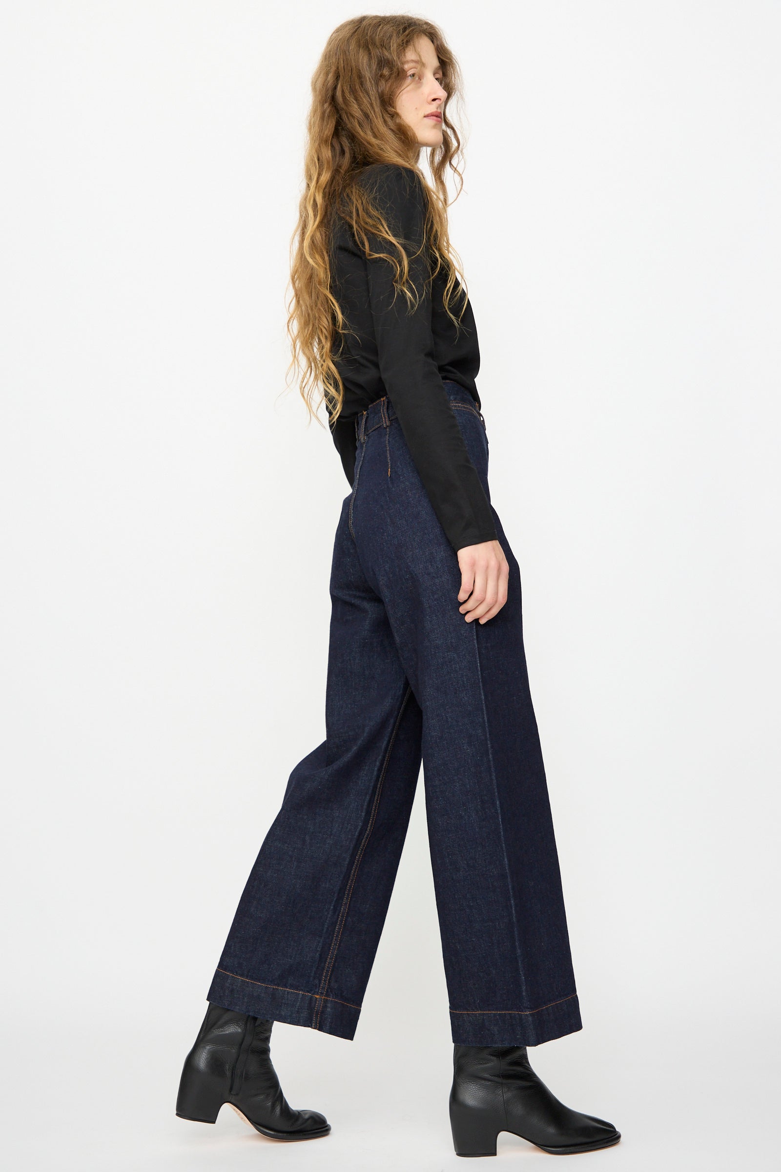 A person with long wavy hair stands sideways against a plain white background, wearing a black long-sleeve top, black heeled boots, and the Jesse Kamm Sailor Pant in Dark Blue.