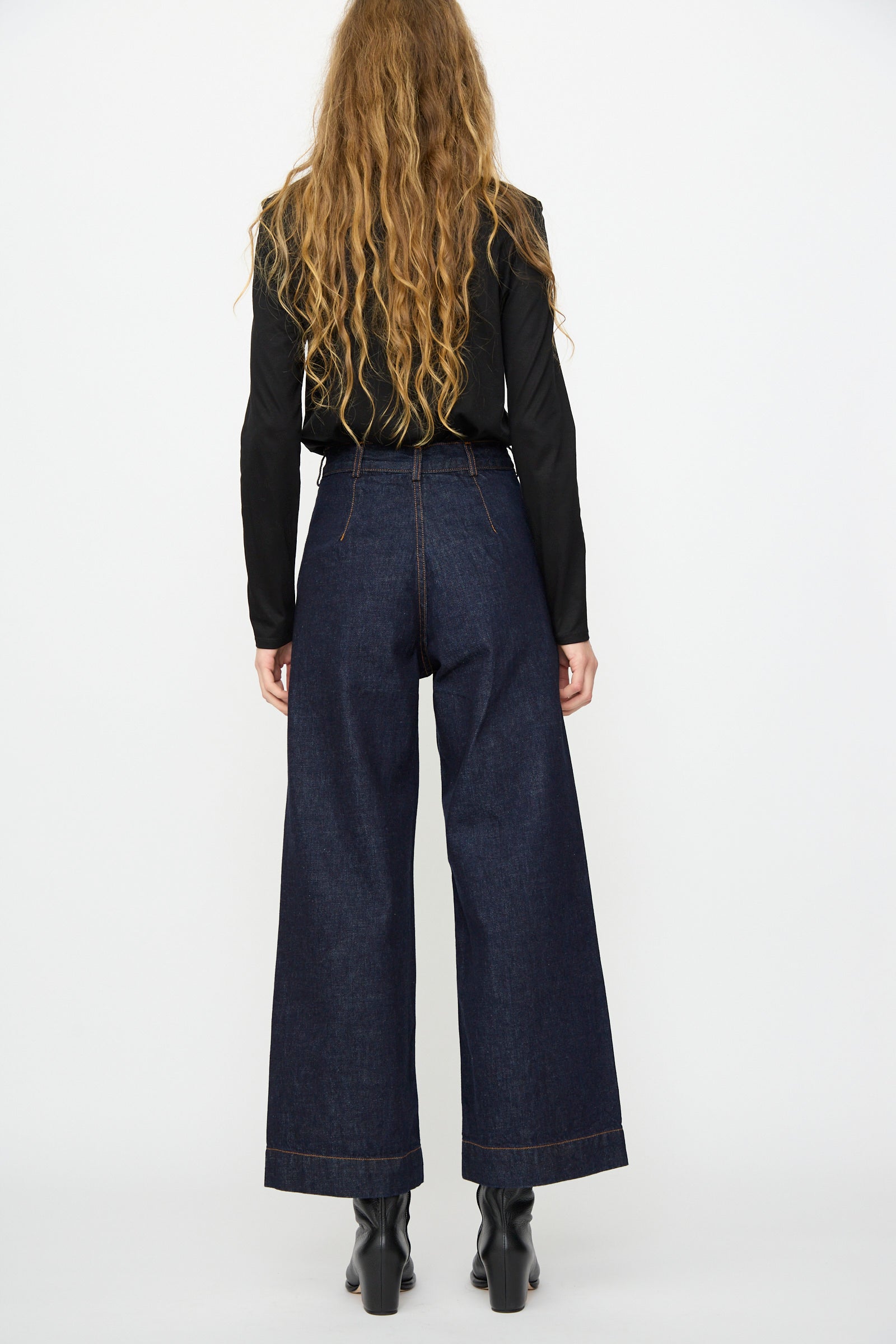 A person with long wavy hair, wearing a black top and Jesse Kamm Sailor Pant in Dark Blue, stands facing away from the camera against a plain white background.