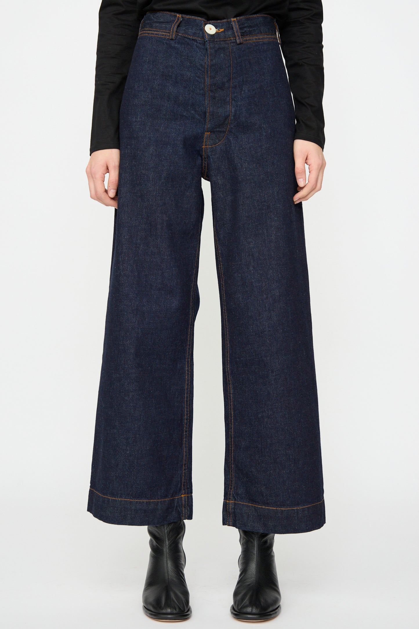 A person wears the Jesse Kamm Sailor Pant in Dark Blue with black boots and a long-sleeve black top, standing against a plain white background.