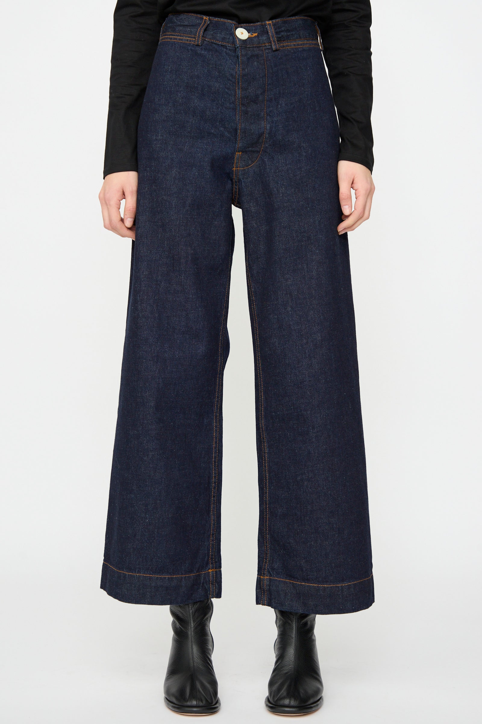 A person wears the Jesse Kamm Sailor Pant in Dark Blue with black boots and a long-sleeve black top, standing against a plain white background.