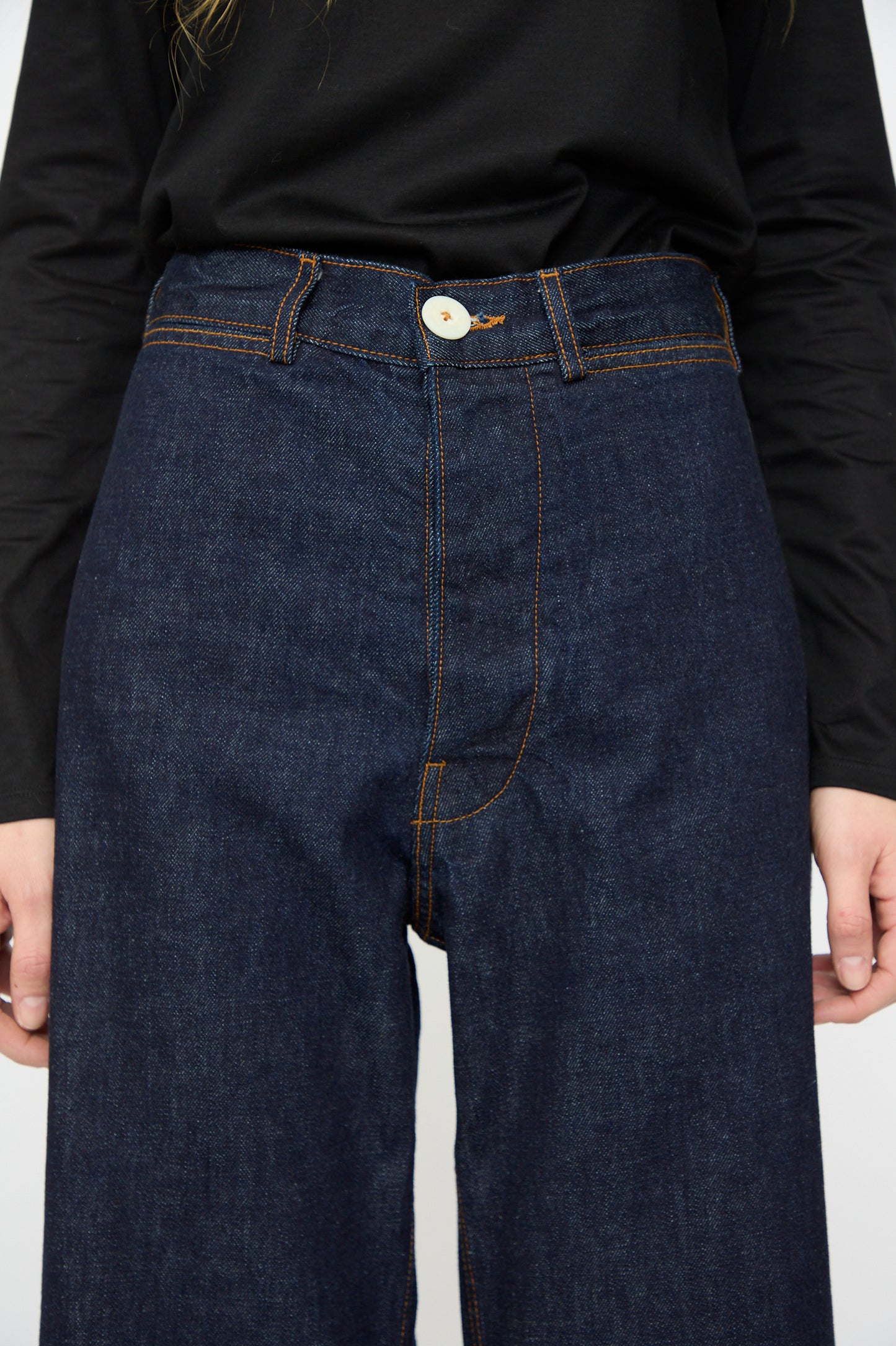 A person wearing the Jesse Kamm Sailor Pant in Dark Blue, featuring high-waisted Japanese denim with orange stitching, paired with a black long-sleeve shirt; image highlights the torso and upper legs.